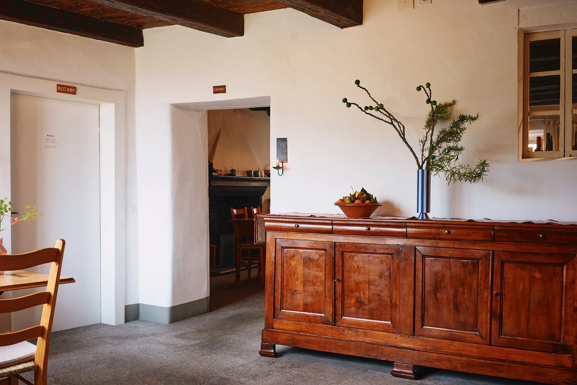 Interno di una casa con pareti bianche e soffitto con travi di legno, arredato con un mobile in legno scuro con vaso di fiori e frutta sopra, e un angolo cucina visibile in una nicchia.
