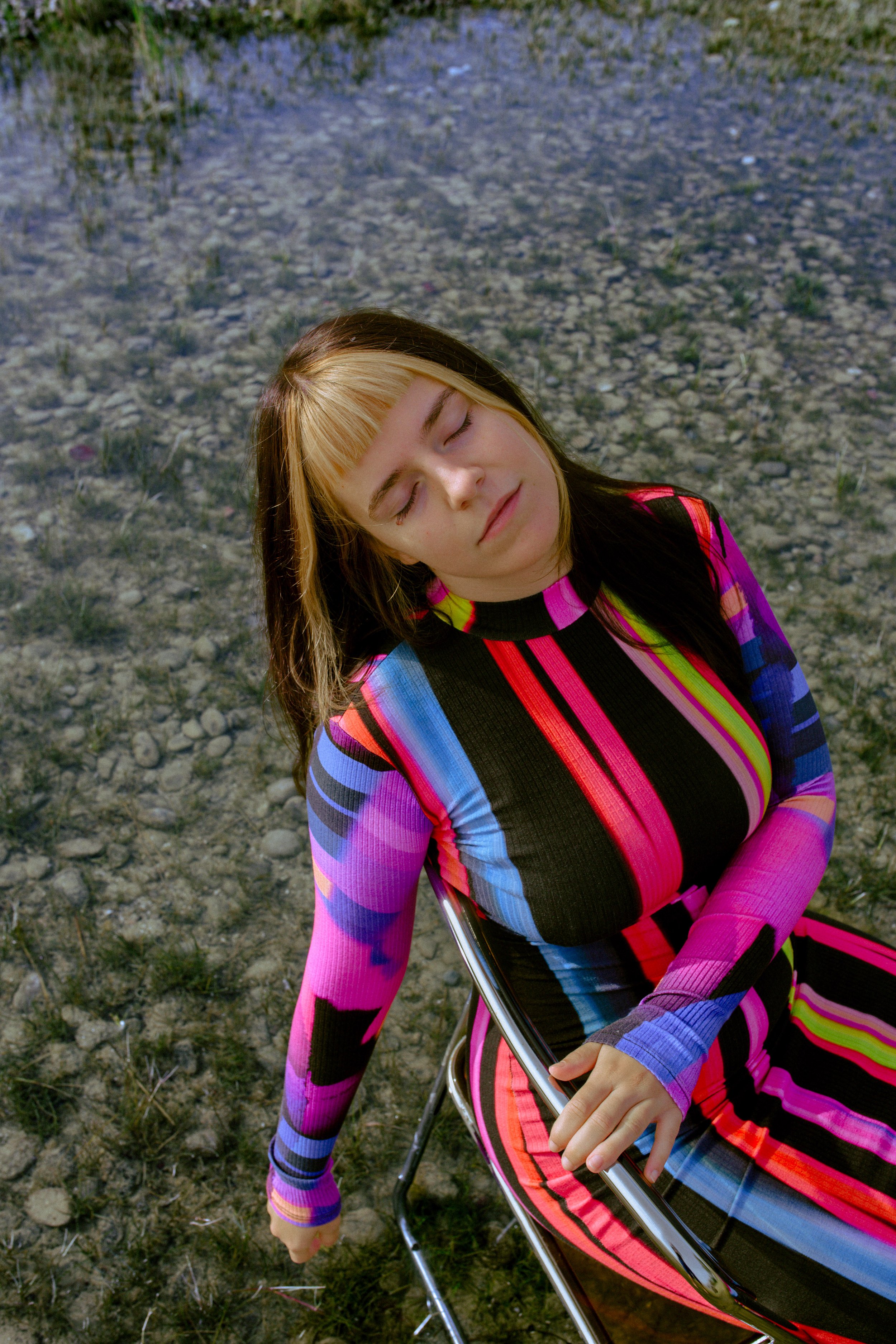 Frau in einem bunt gestreiften Kleid sitzt entspannt mit geschlossenen Augen im Freien auf einem Stuhl.