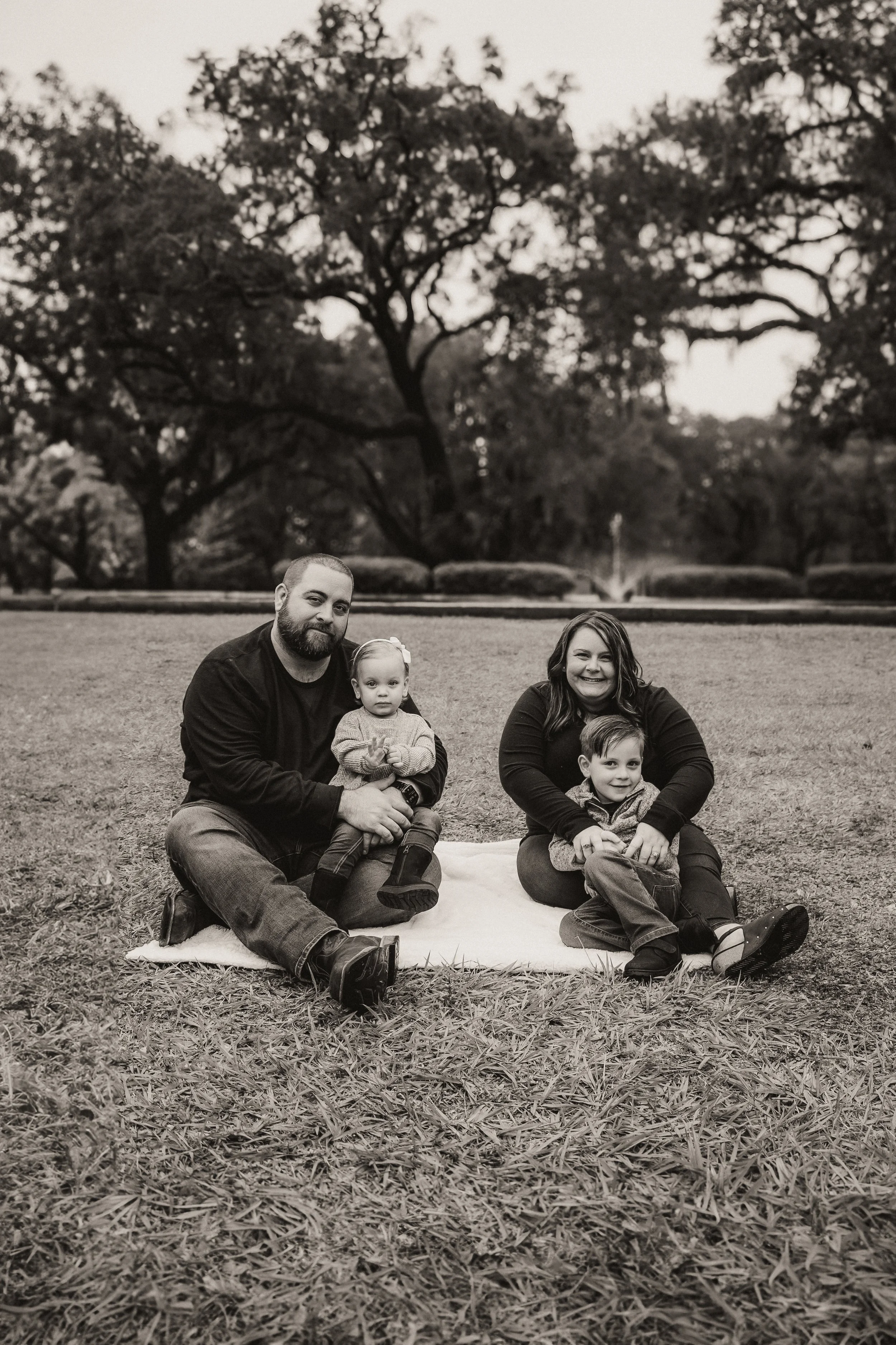 Black and white photograph of a family of four sitting outdoors on a blanket in a park. Trees and bushes are visible in the background.