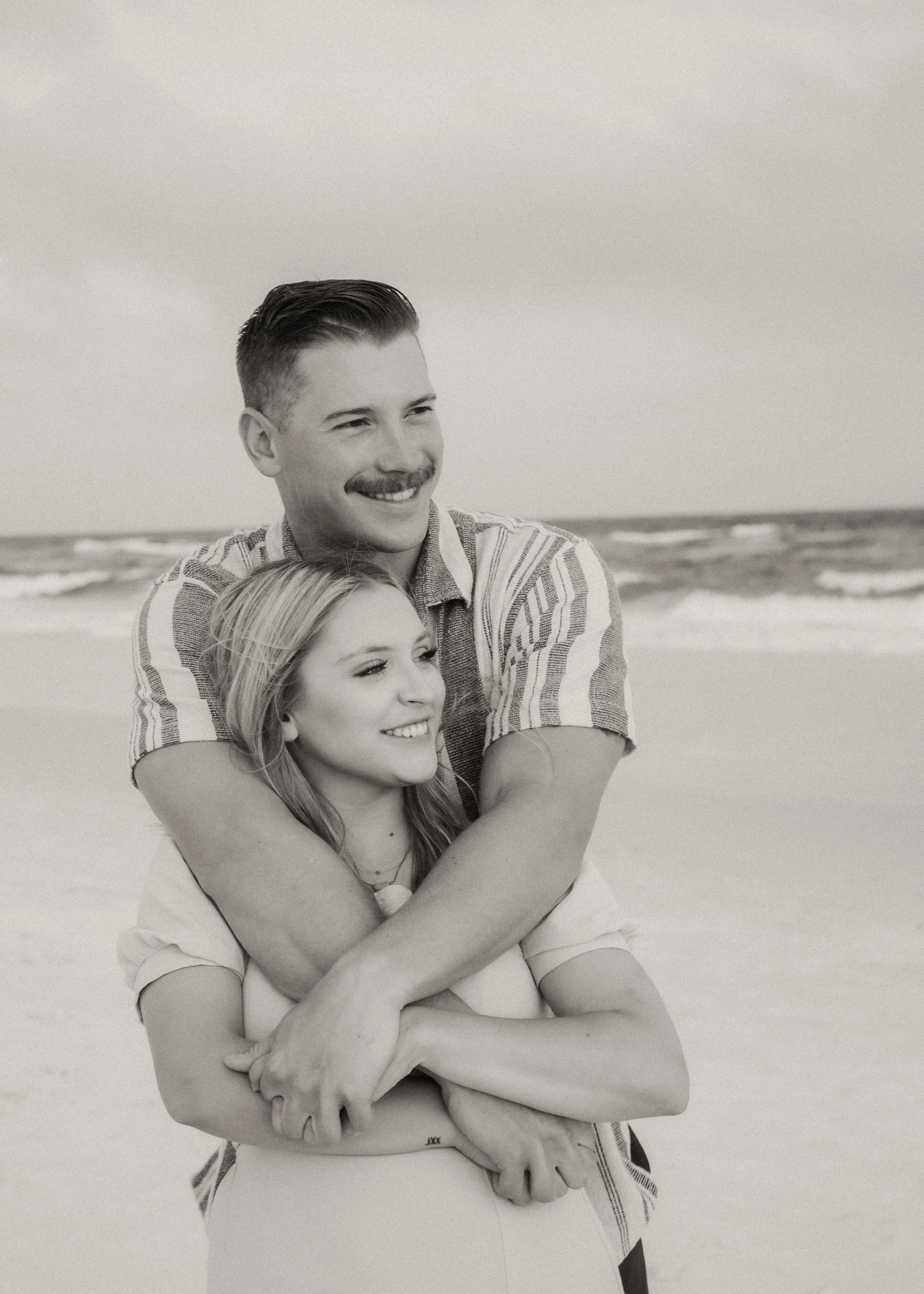 A black and white photo of a smiling couple on the beach, with the man hugging the woman from behind. The ocean and sky are visible in the background.