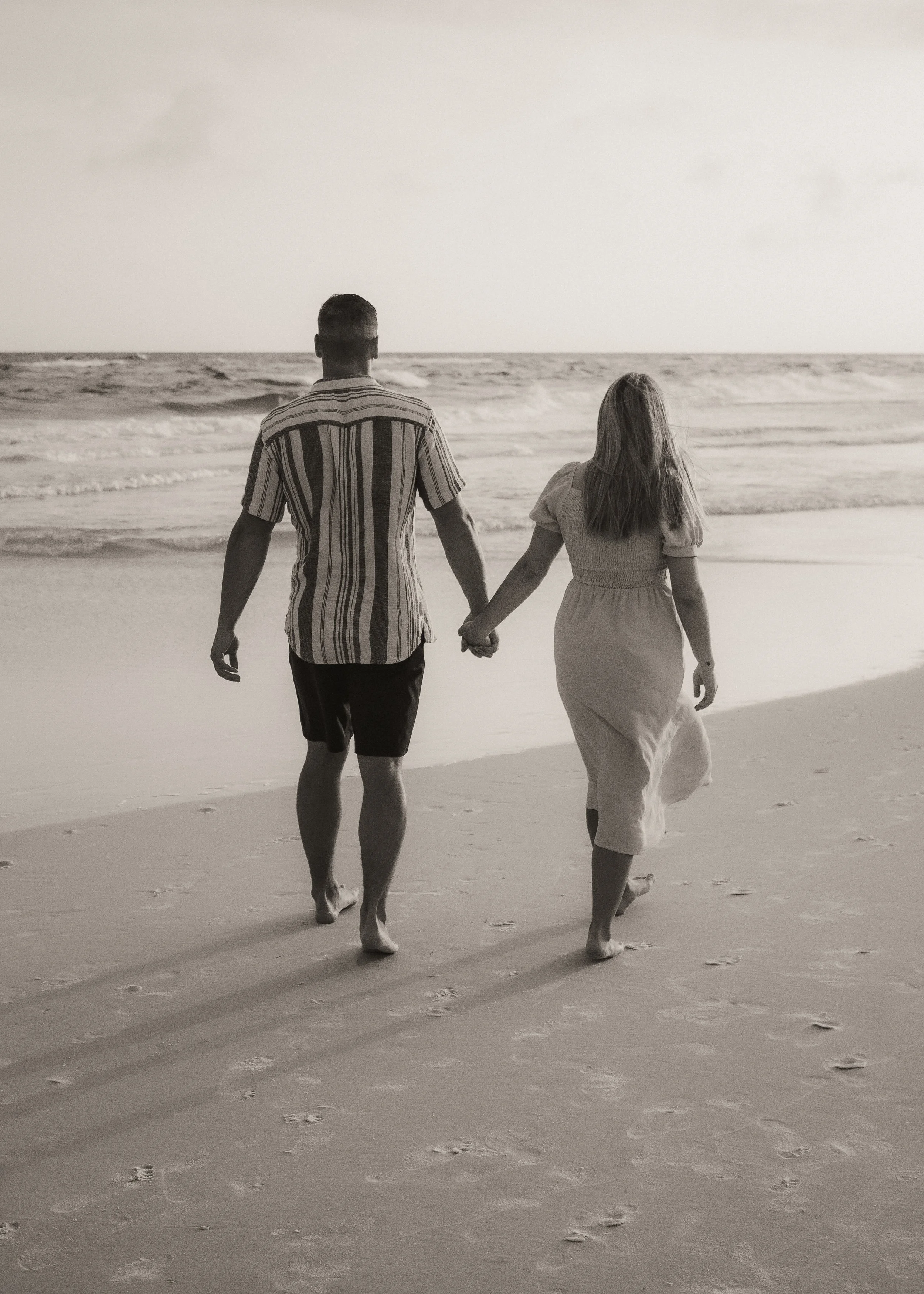 A black-and-white photo of a couple walking hand in hand on the beach, facing the ocean.