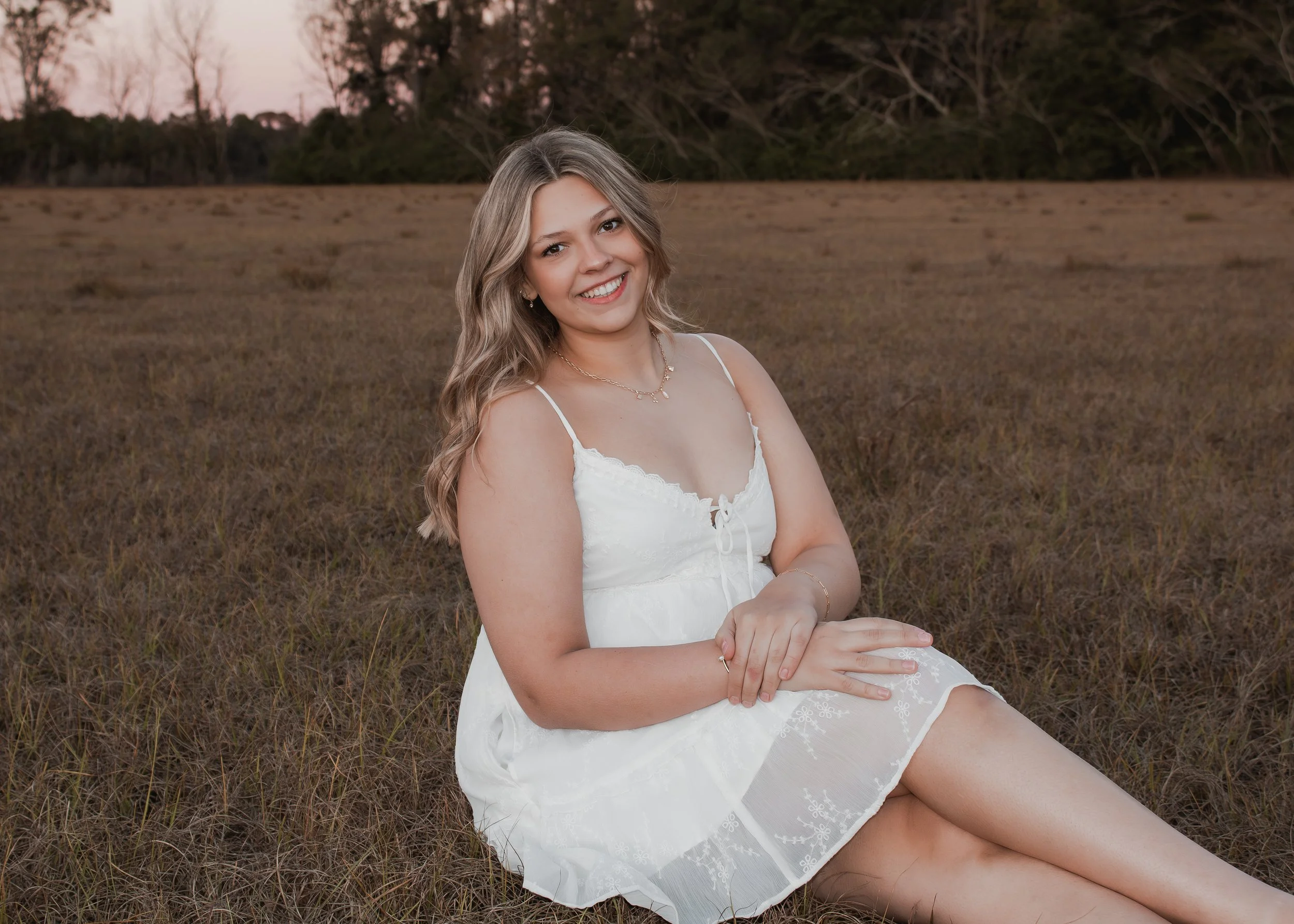 A young woman with blonde, wavy hair smiling and sitting on grass in a field at sunset.