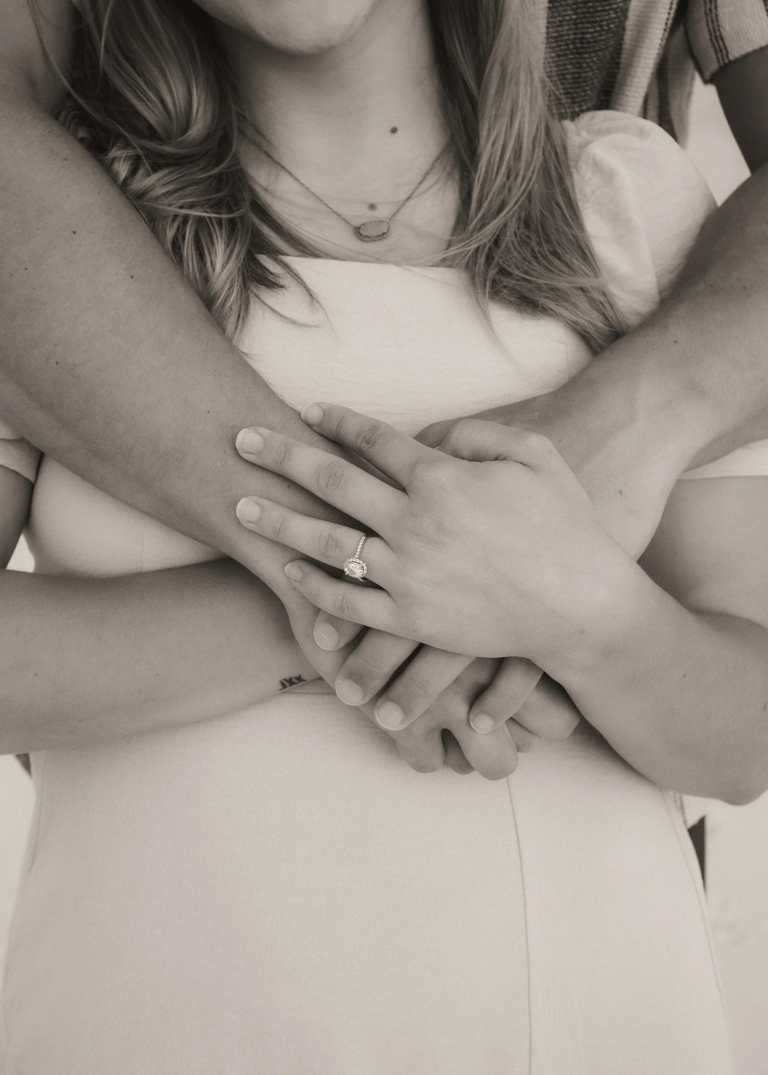 Close-up of four hands gently holding, two of which are intertwined, with a woman showing a wedding ring on her finger. The woman has wavy hair, wears necklaces, and a light-colored top.