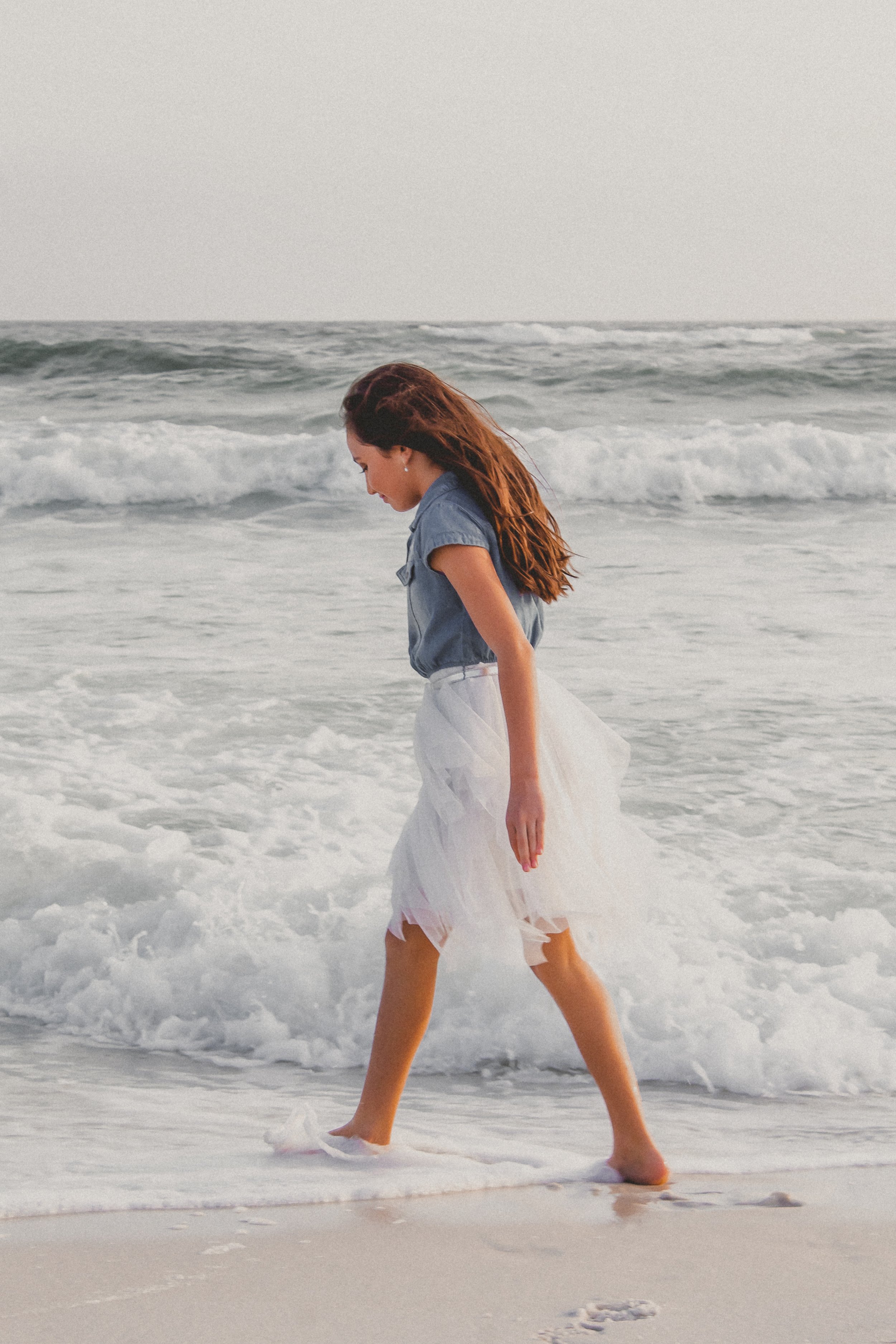 A woman with long brown hair walks along the shore of the ocean, wearing a blue shirt and a white skirt, with waves washing over her feet.