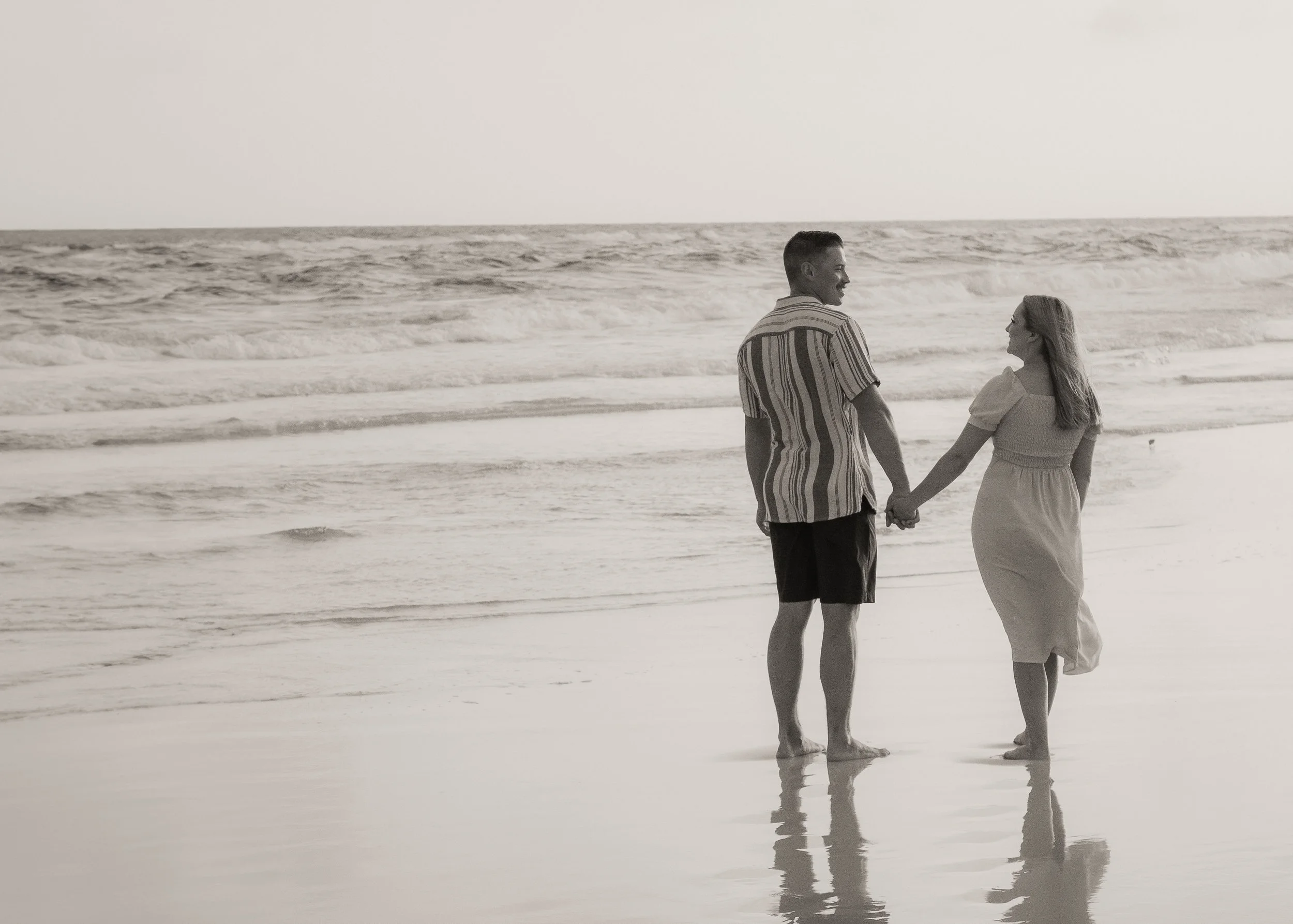 A couple holding hands on the beach, walking near the water, in black and white.