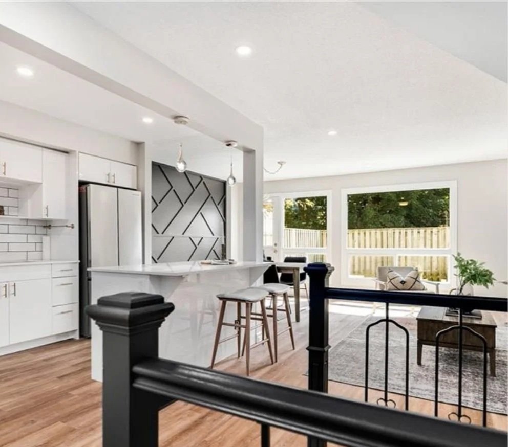 Interior view of a modern, bright living space with white cabinets, a stainless steel refrigerator, a kitchen island with bar stools, and large windows with a view of a green backyard with a wooden fence.