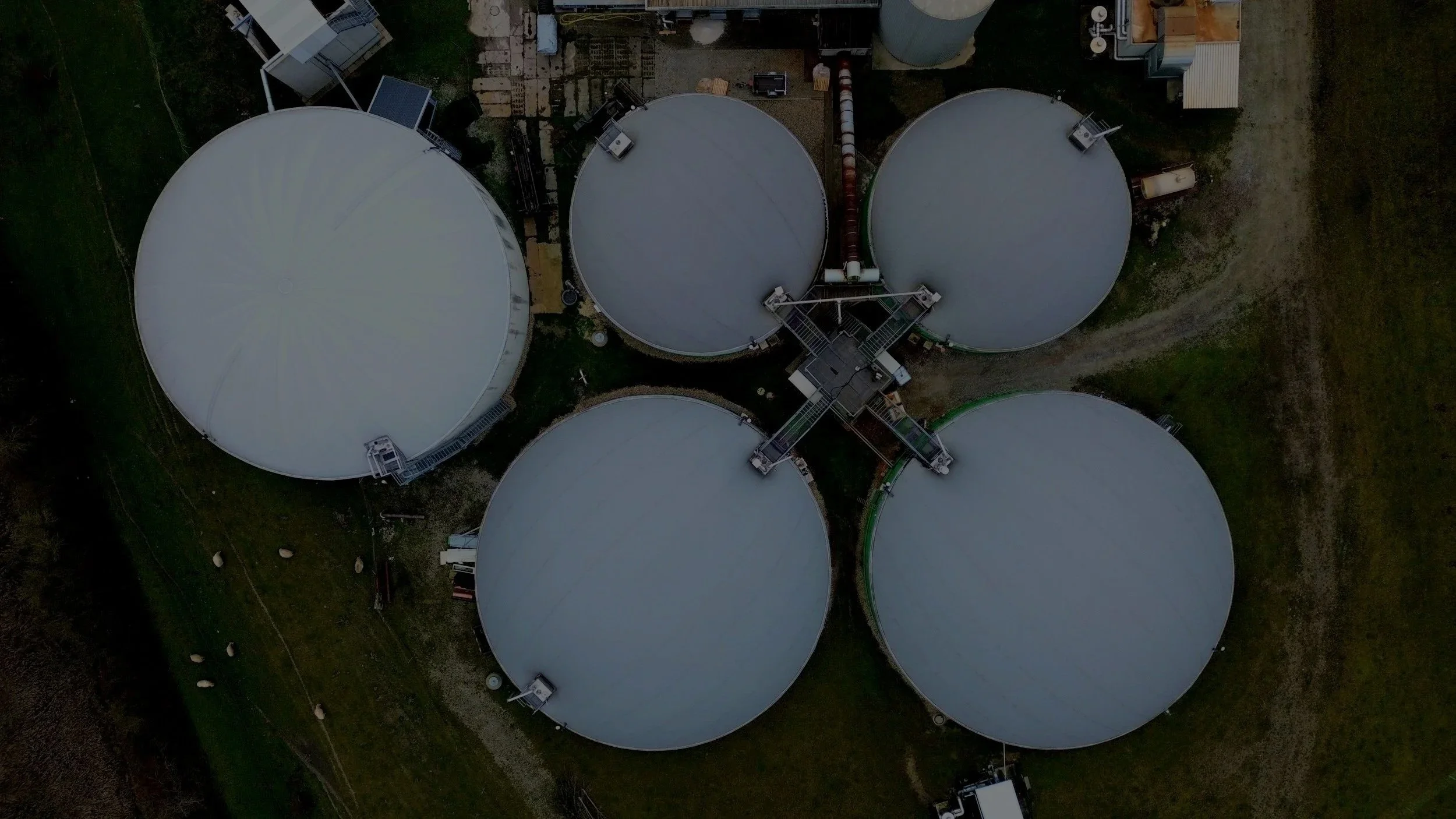 An aerial view of a facility with five large circular storage tanks arranged in a cluster, connected by walkways and pipelines, surrounded by grass and dirt paths.