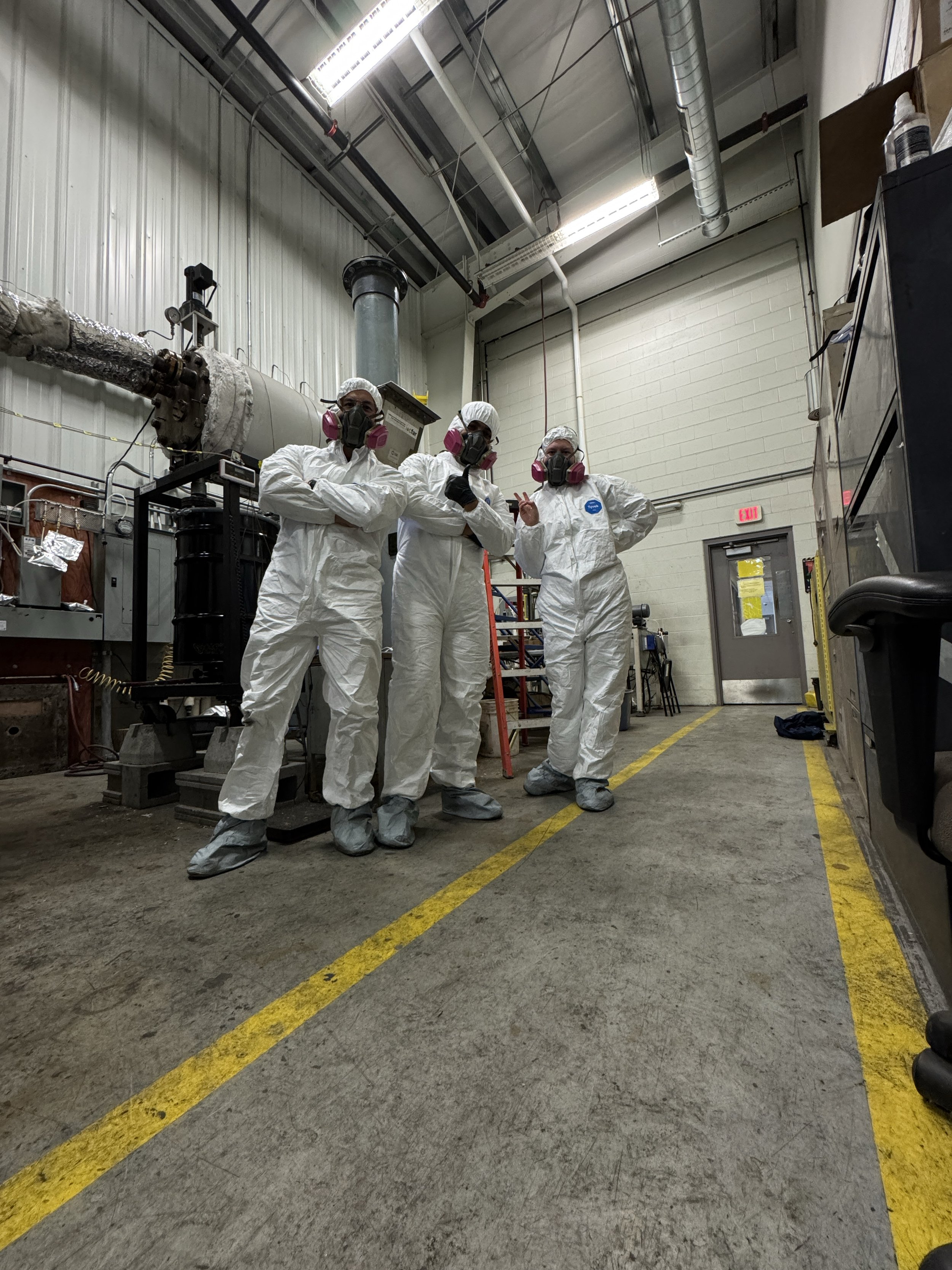 Three people dressed in white protective suits, masks, and gloves standing in a laboratory or industrial setting, posing for a photo.