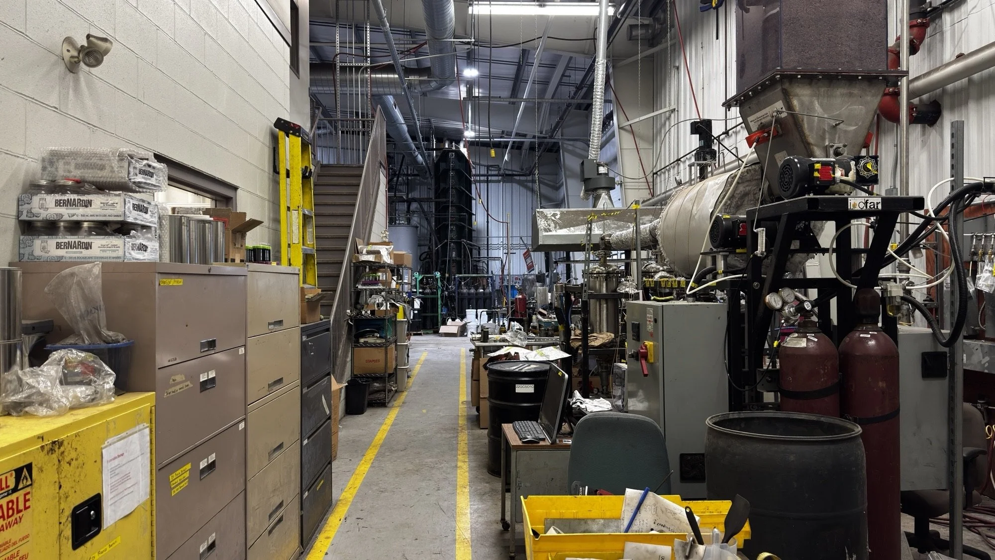 An industrial laboratory workspace with various equipment, tools, and storage cabinets. There are pipes and tanks, a yellow ladder, a computer on a desk, and cluttered shelves.