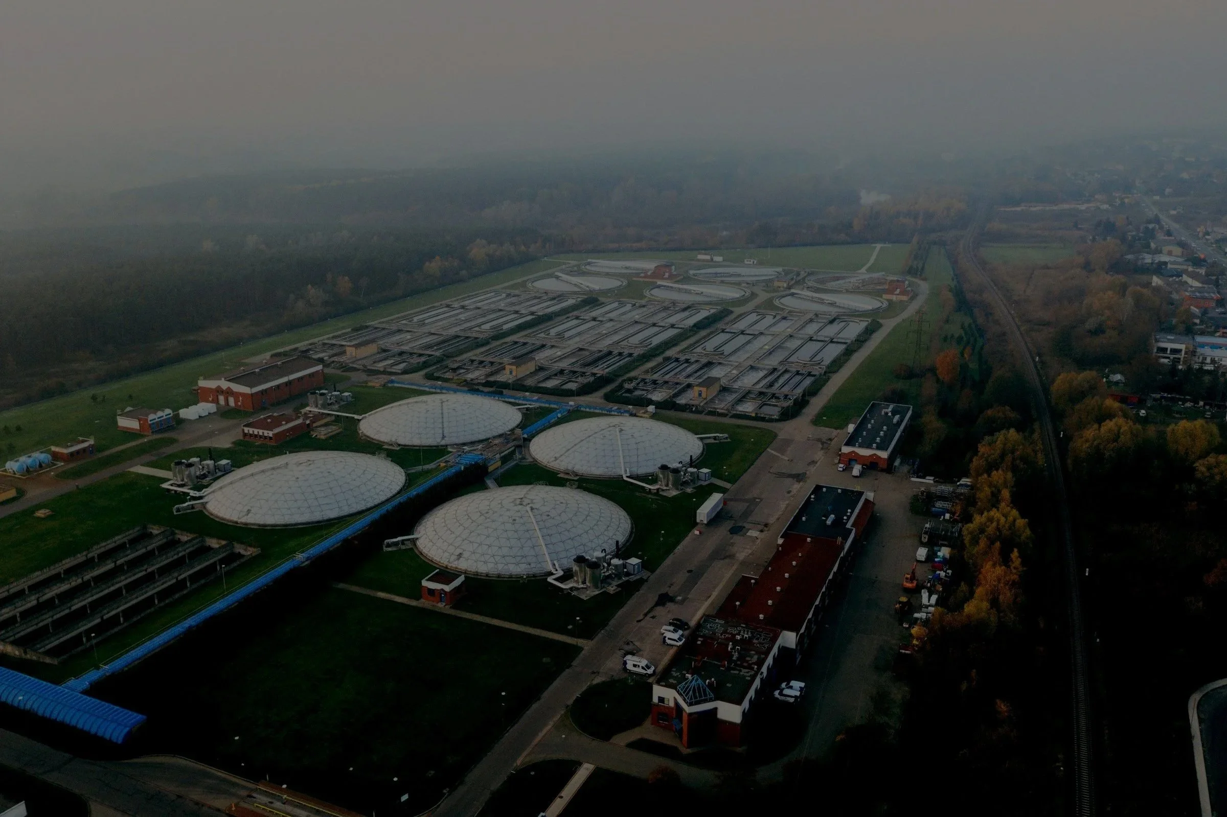 Aerial view of a water treatment plant with multiple large white domed structures, rectangular buildings, water tanks, and surrounding infrastructure, with trees and a railway track to the right.