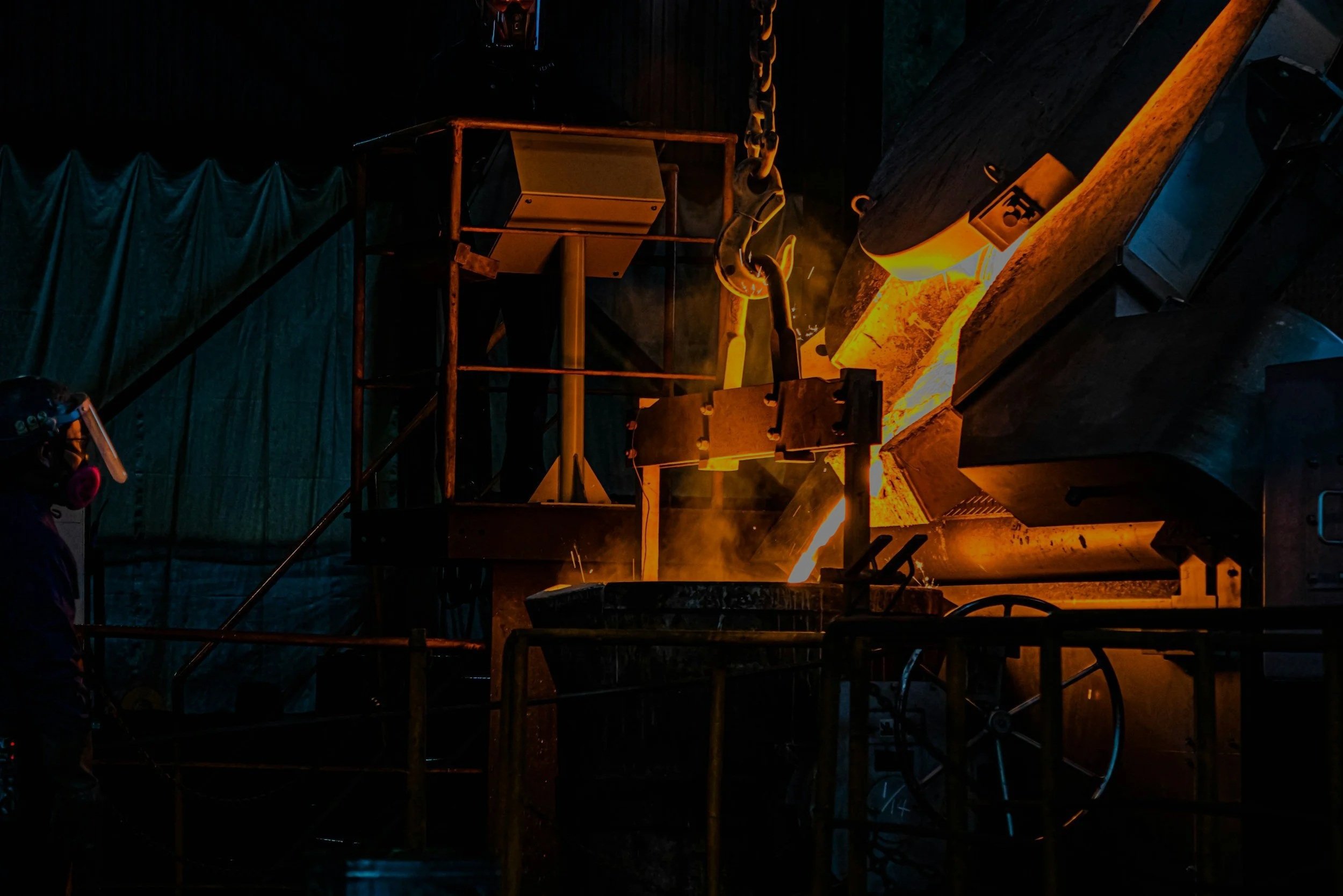 Industrial worker wearing protective gear and a headset operating heavy machinery in a foundry, melting metal with bright orange flames and glowing hot metal.