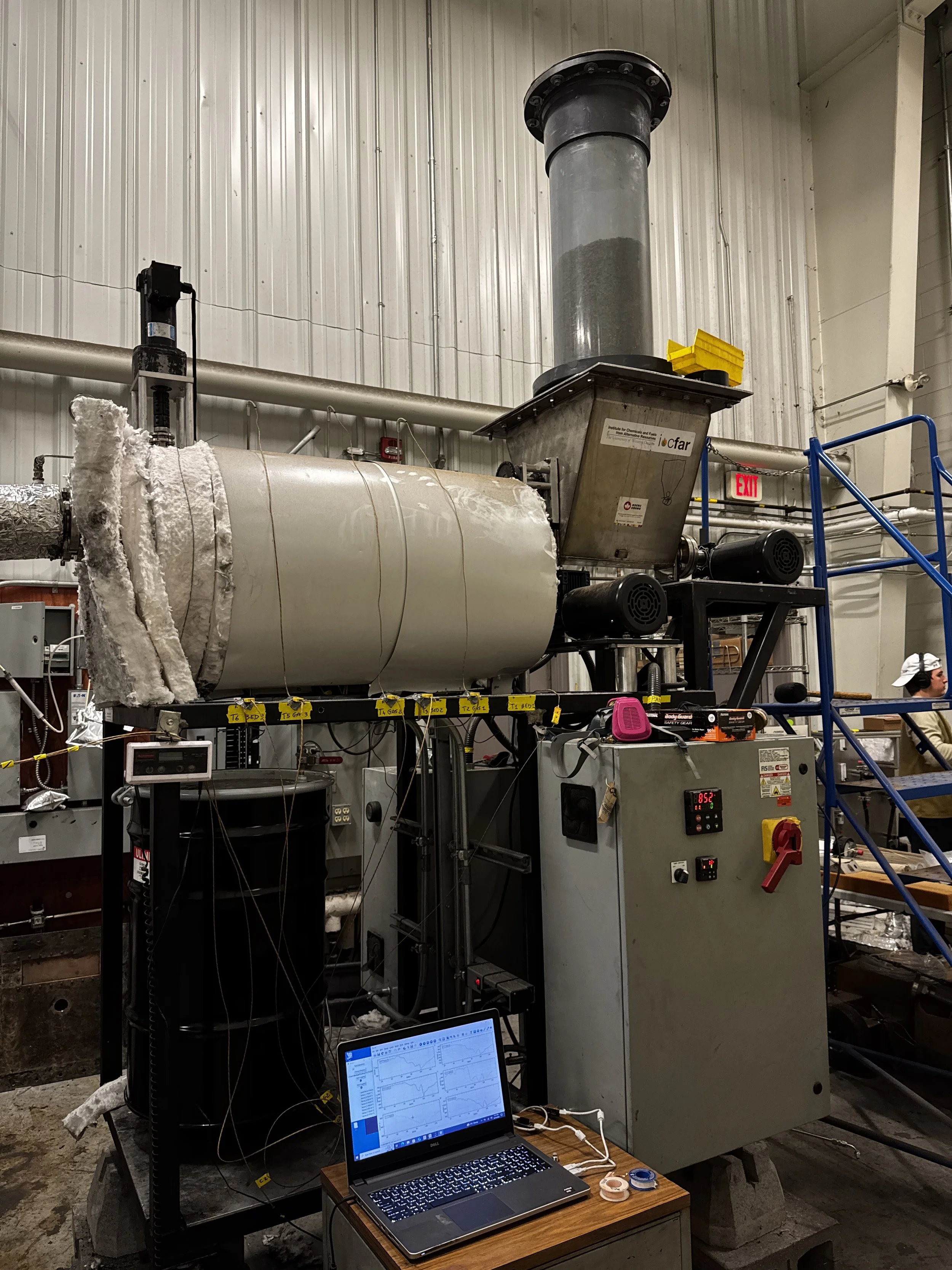 Industrial laboratory setup with large cylindrical reactor vessel, control panel, laptop displaying graphs, and various equipment in a warehouse setting.