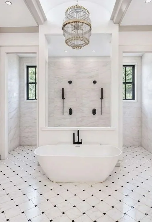 Luxury bathroom with white freestanding bathtub, black fixtures, marble walls, black and white tiled floor, two windows, and a chandelier.