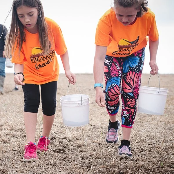 Two young girls wearing orange t-shirts and colorful leggings or pants, each carrying a white bucket, walking on a grassy or sandy outdoor area near a body of water.