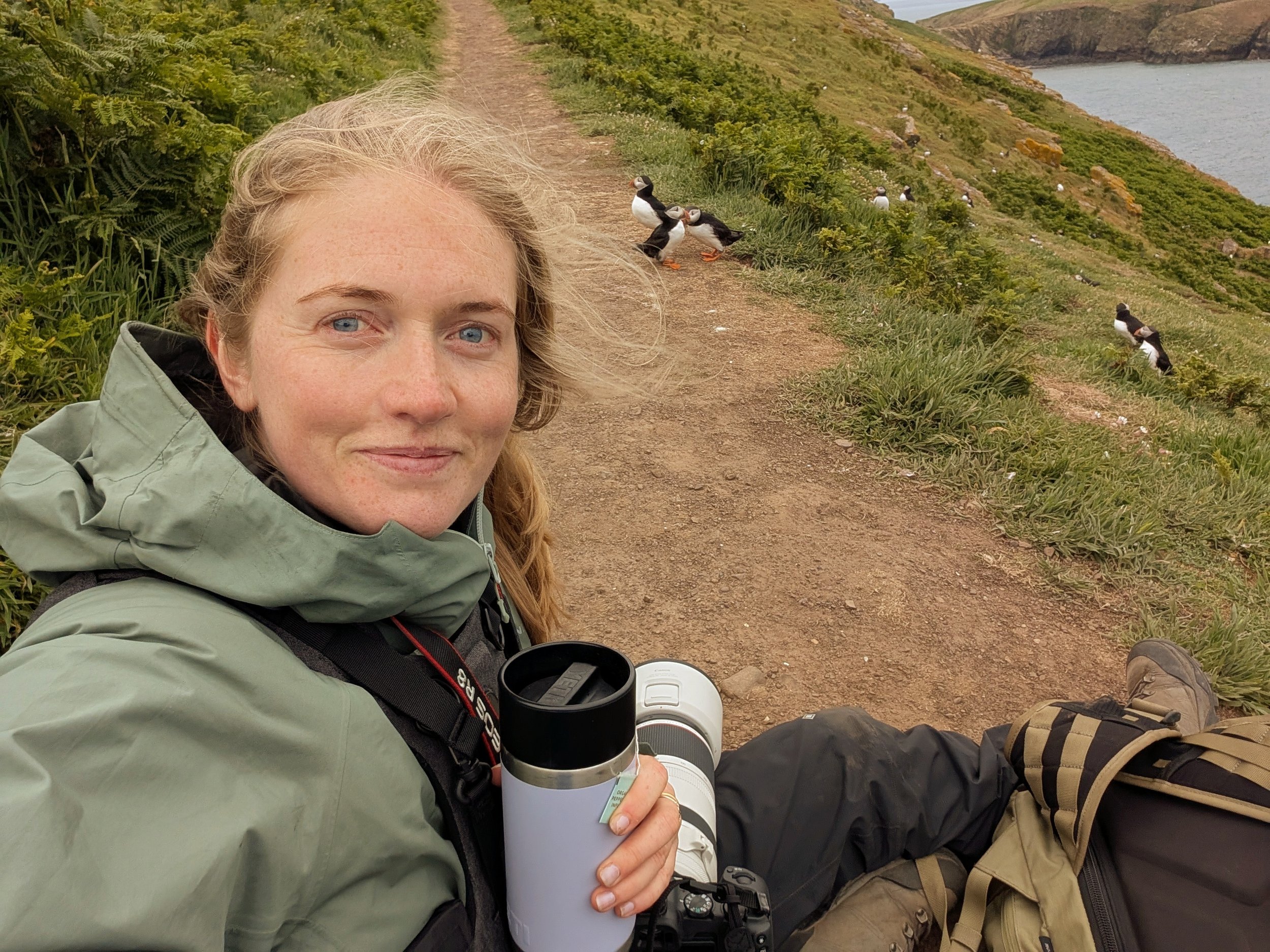 A woman with long red hair and blue eyes sitting on a dirt trail near the coast, holding a white travel mug, with penguins and seagulls nearby on the grassy slope.