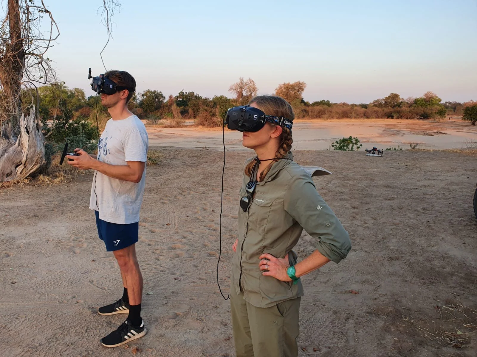 Two people wearing virtual reality headsets stand outdoors in a sandy, dry area with sparse trees. One person is a woman with a braid, wearing a beige jacket and pants, and the other is a man with short hair, wearing a light t-shirt and shorts. The b