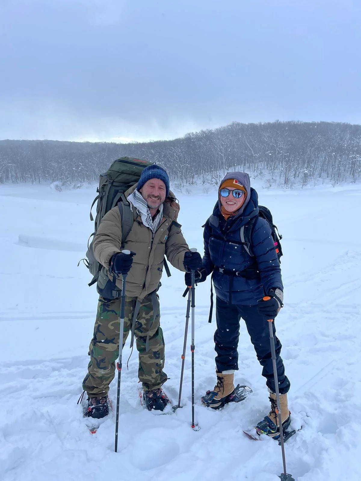 Two people in winter clothing and snow boots standing on snow, holding trekking poles and smiling, with a snowy landscape and trees in the background.
