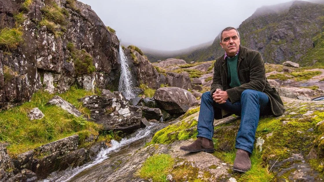 A man sitting on a moss-covered rock with a small waterfall and rugged, mossy mountain landscape in the background.