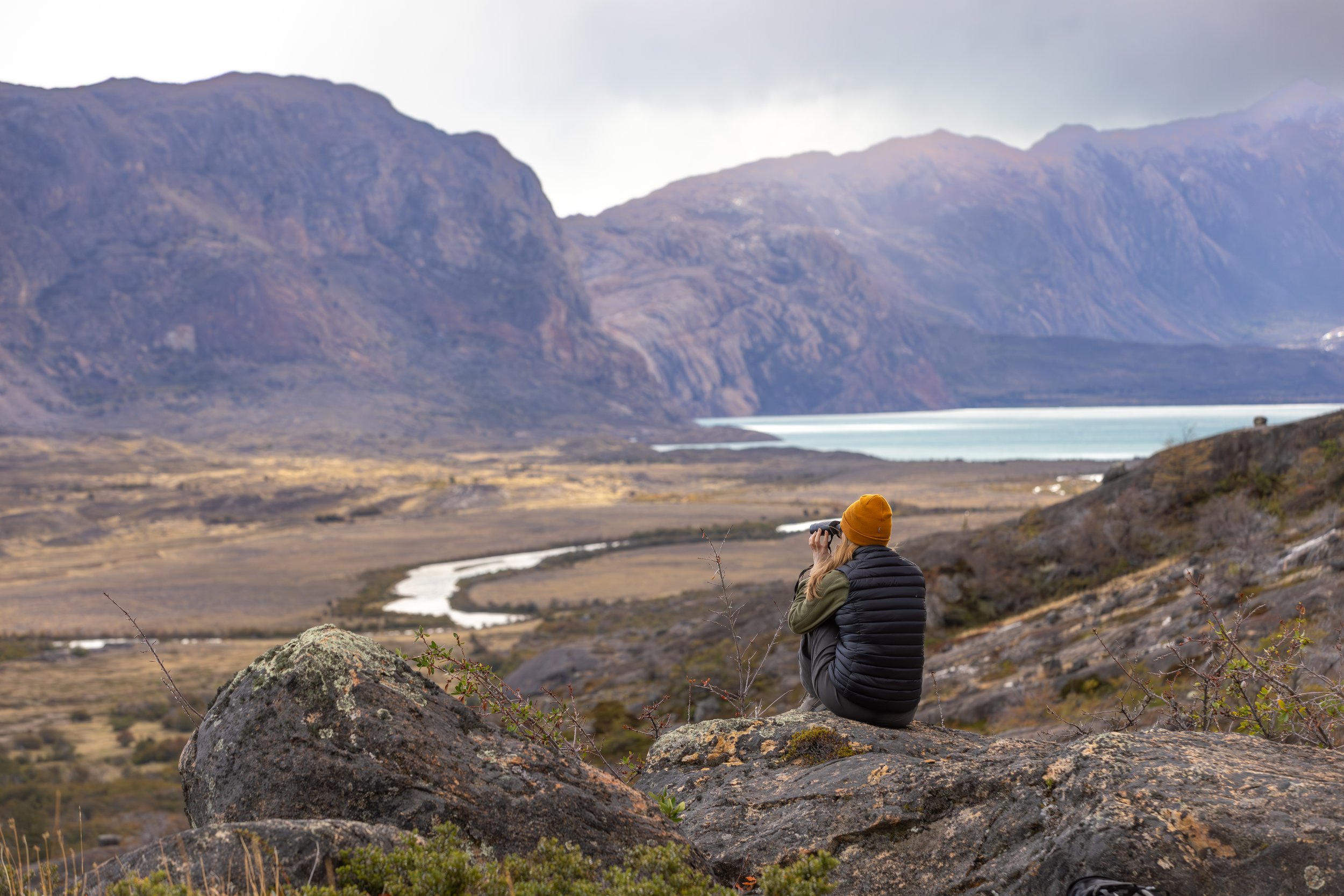 Person sitting on rocks in a mountainous landscape taking a photo with binoculars, wearing a yellow beanie and a black vest.