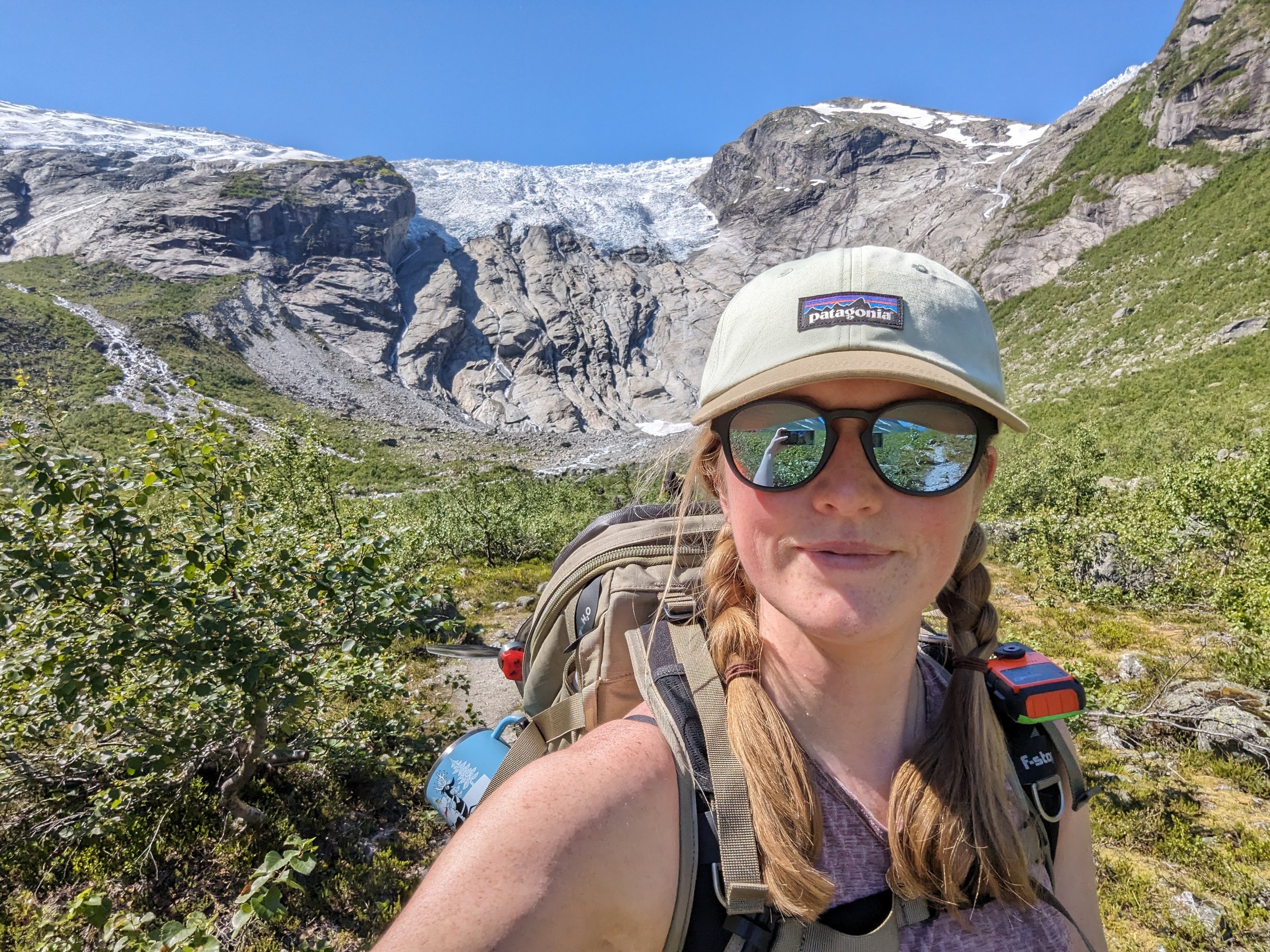 Woman hiking in a mountainous area with snow-capped peaks, wearing a beige cap, sunglasses, and a backpack.