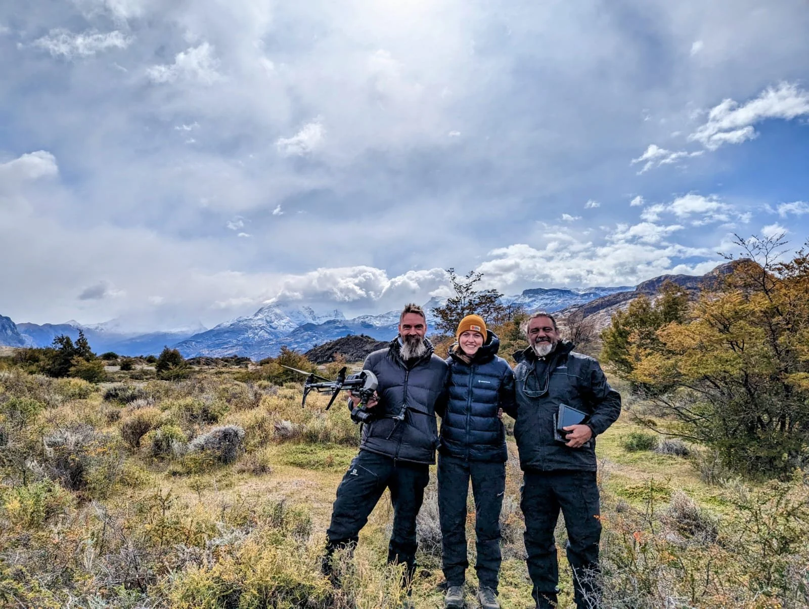 Three people standing outdoors in a mountainous landscape, smiling and posing for the photo; they are wearing outdoor gear, with one holding a drone, and the background shows trees, mountains, and a partly cloudy sky.