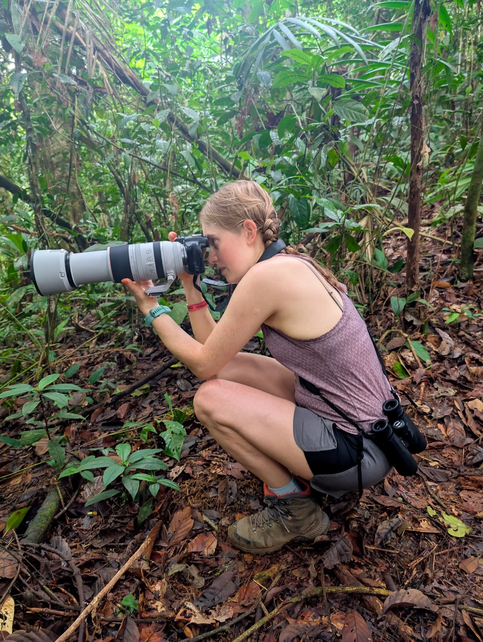 Briony Jones in the jungle in Costa Rica, looking through a large camera with a telephoto lens, wearing hiking boots, shorts, and a sleeveless top, with binoculars hanging from her waist.