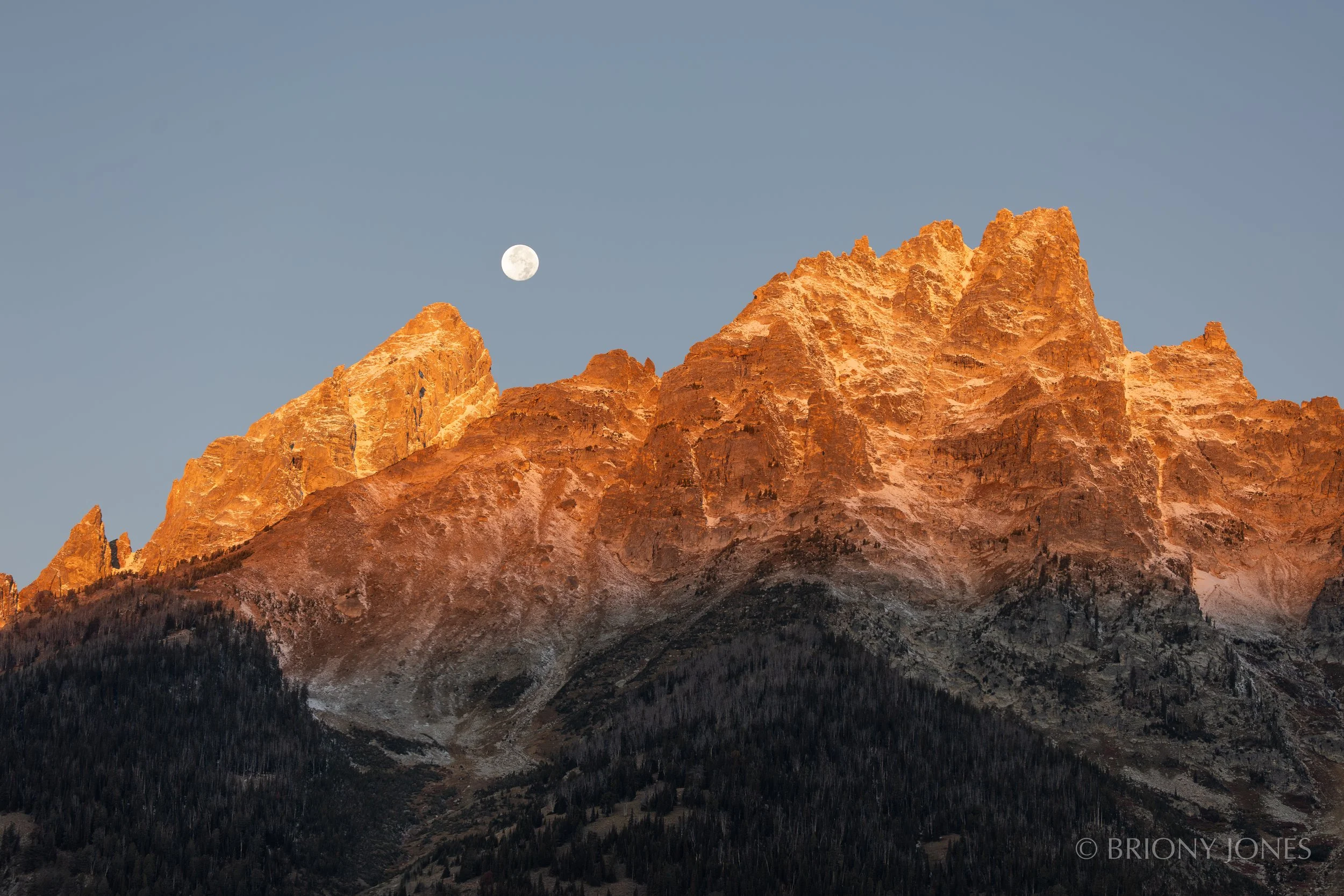 Mountain range with rocky peaks illuminated by the setting sun and the moon visible in the sky.