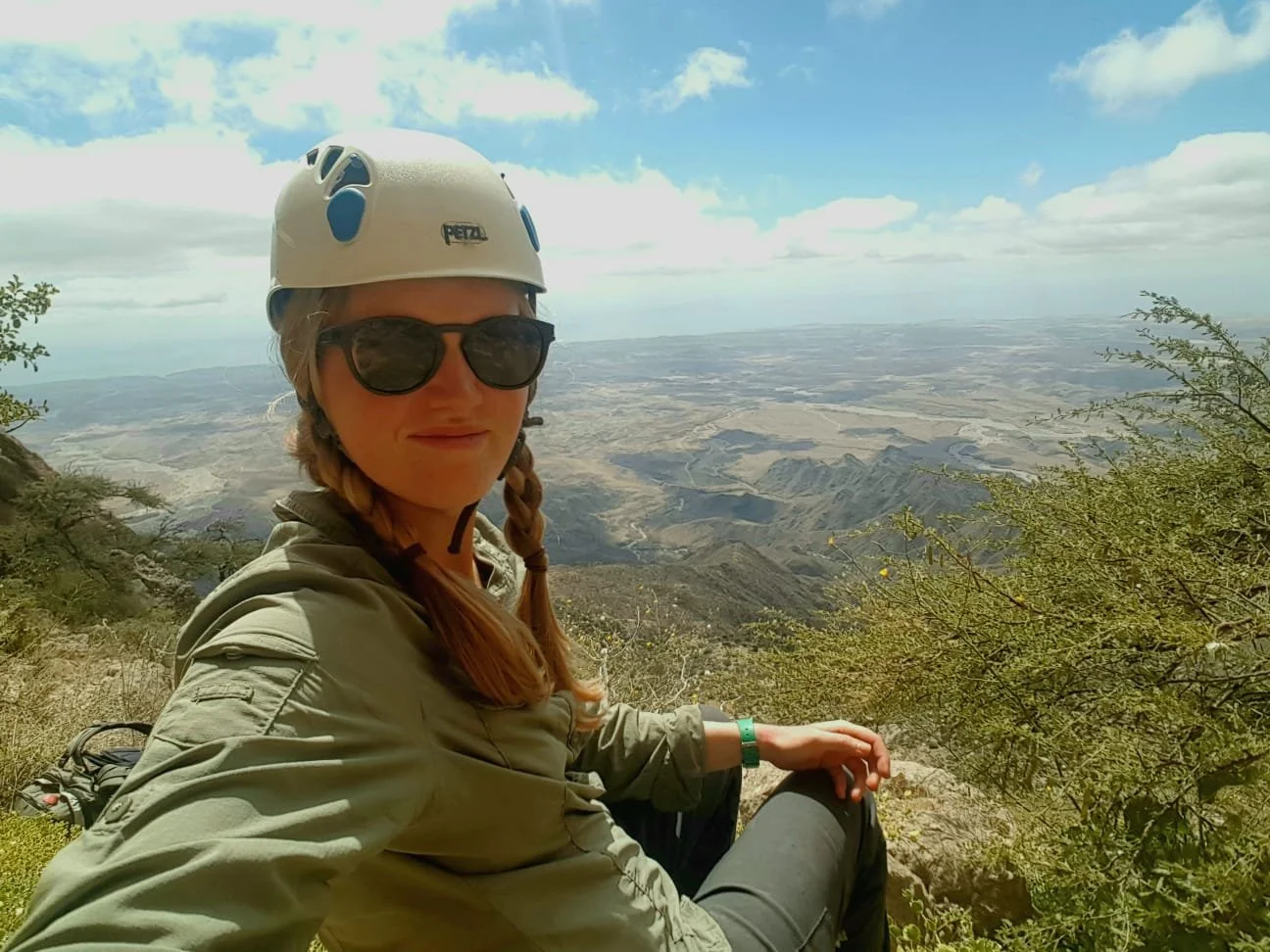 A woman wearing a white climbing helmet and sunglasses sitting on a hillside with a vast desert landscape in the background under a partly cloudy sky.