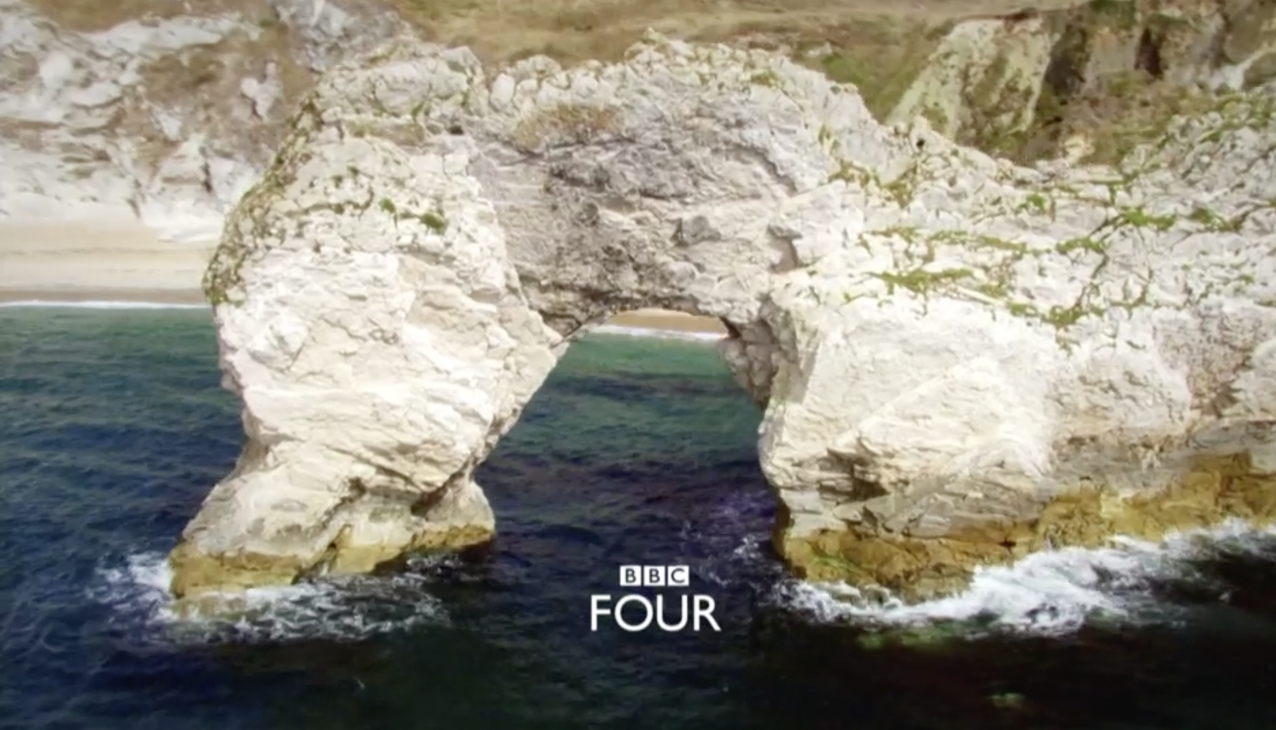 Natural rock arch formation over the ocean with a sandy beach and cliffs in the background, BBC Four logo at the bottom.