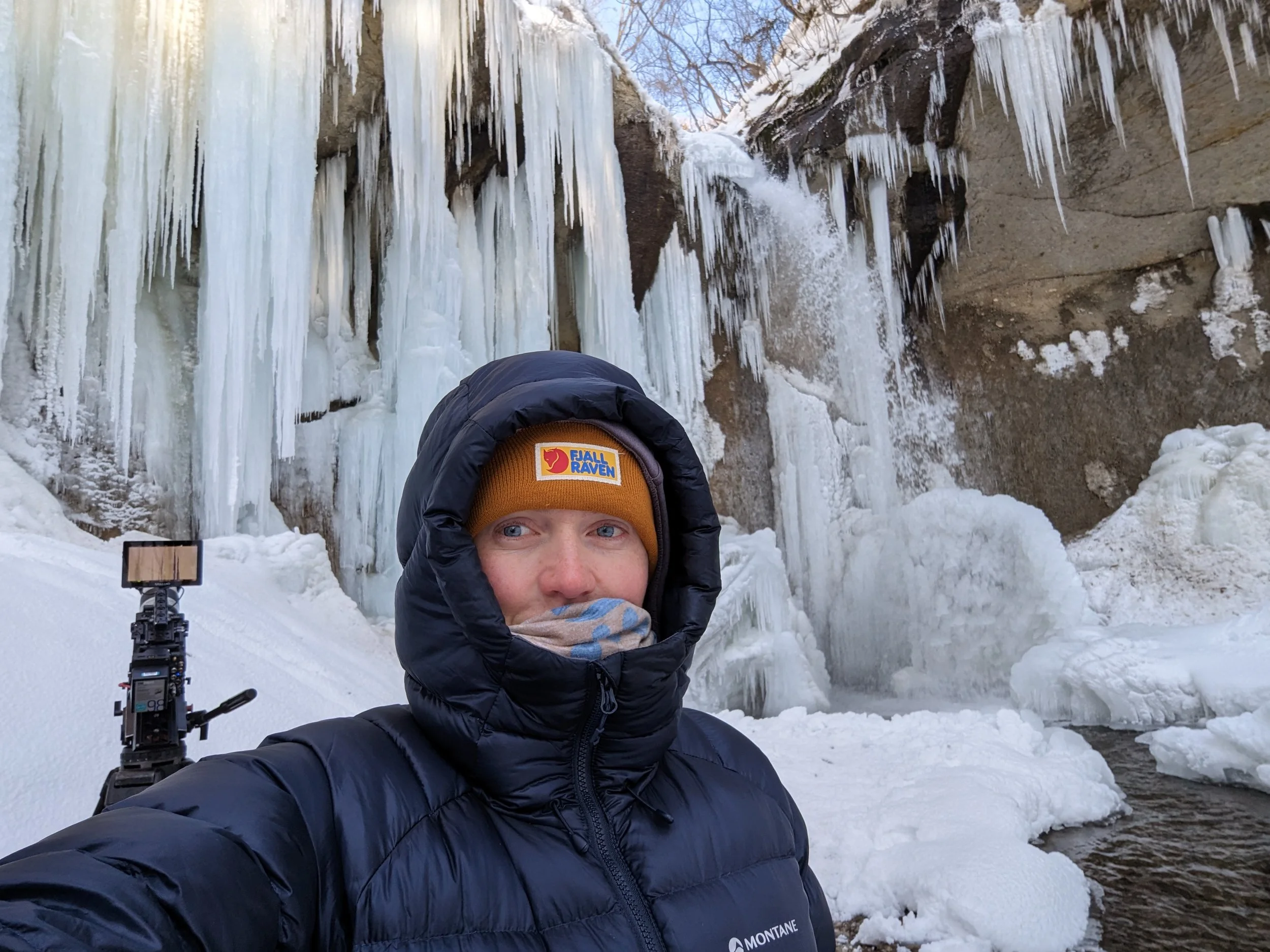 Person taking a selfie in winter clothing stands in front of a frozen waterfall with icicles and snow.