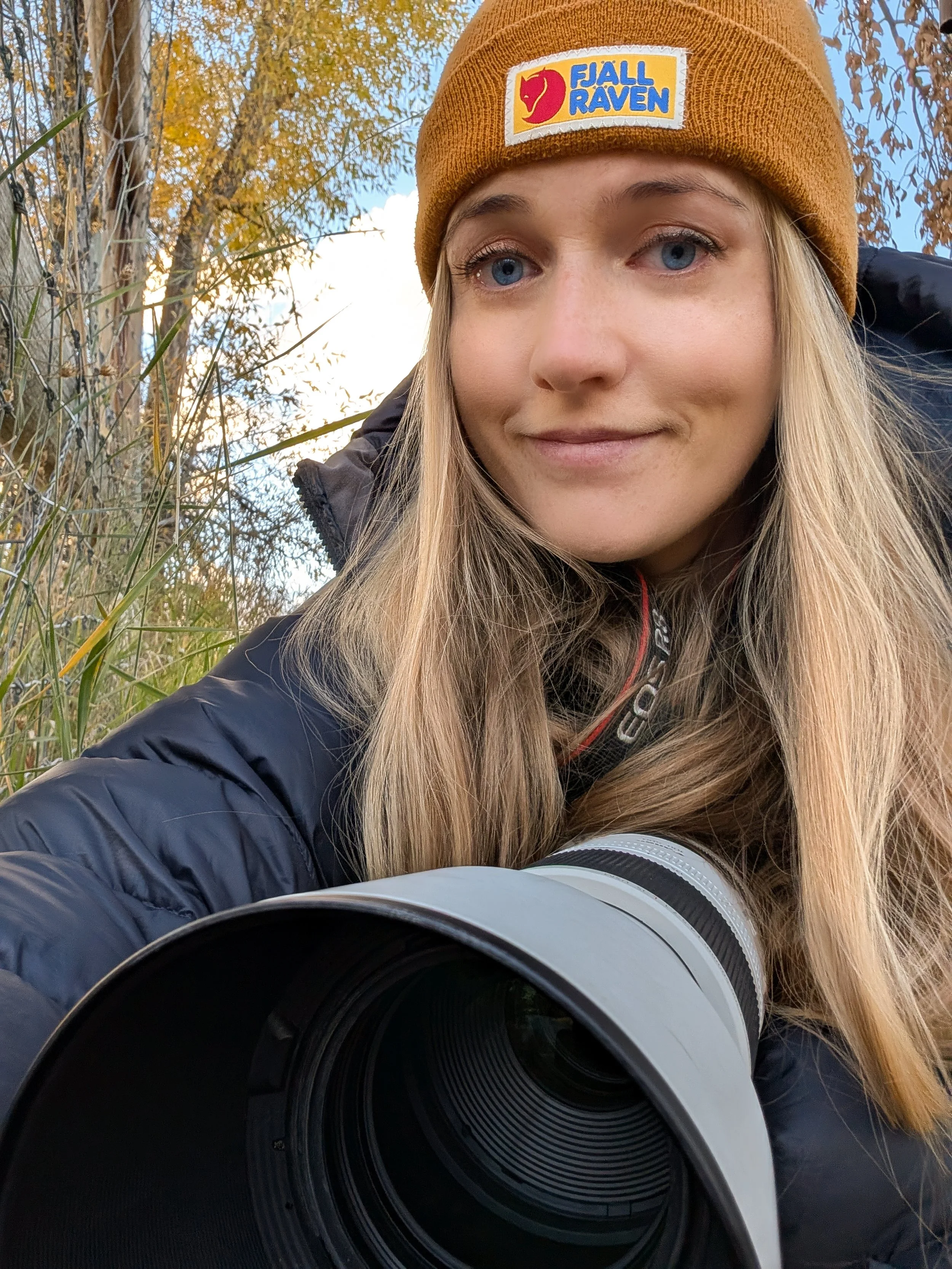 A woman with long blonde hair wearing a yellow beanie with a Fjallraven logo, outdoors in fall, with trees and a camera lens visible in the foreground.