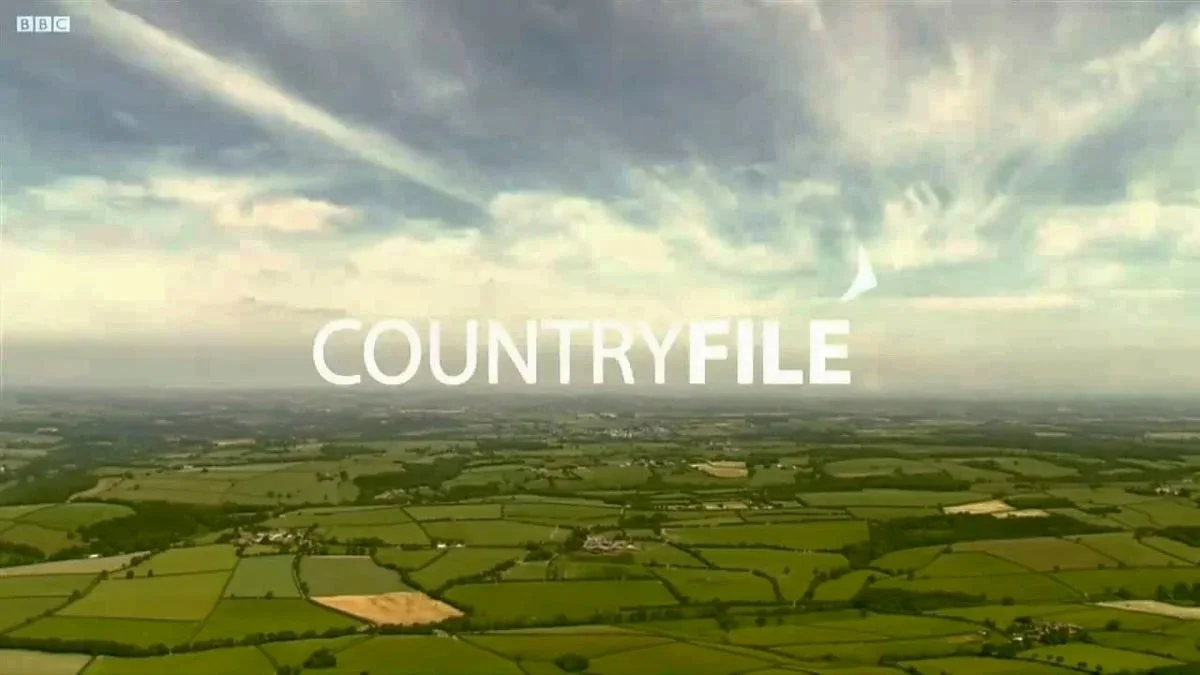 A vast green countryside landscape with fields, hedgerows, and a few scattered buildings, under a partly cloudy sky with the word "COUNTRYFILE" in large white letters across the center.