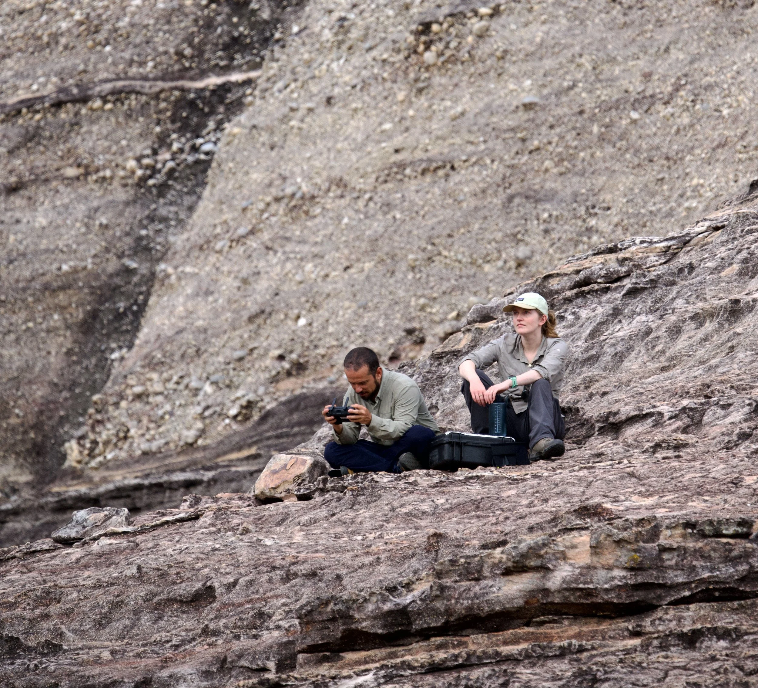 Two explorers sitting on rocky terrain in a desert landscape. One is looking at equipment while the other is gazing into the distance.