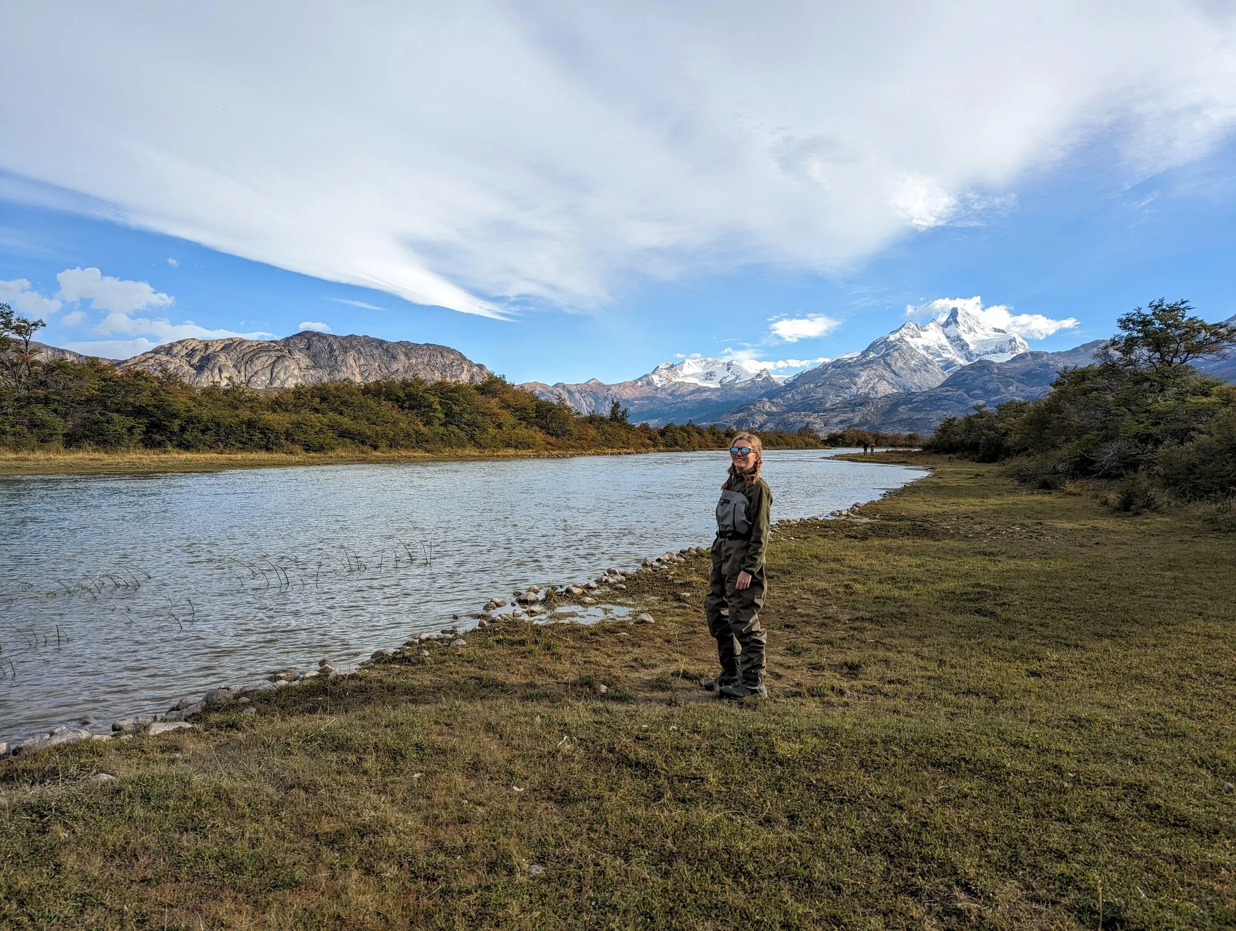 Person standing on grassy riverbank with mountains and snow-capped peaks in the background under a partly cloudy sky.