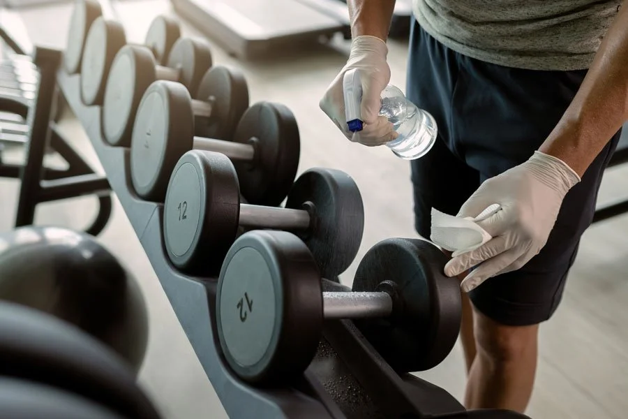 Una persona che disinfetta i manubri in palestra con una bottiglia d'acqua e un panno.