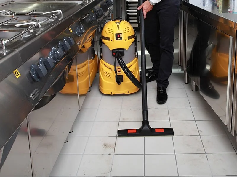 Persona che usa un aspirapolvere industriale in una cucina commerciale con pavimento di piastrelle.