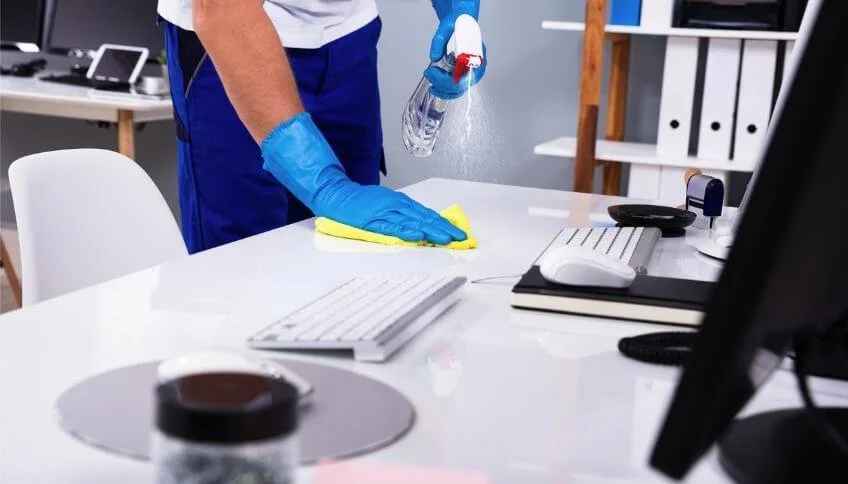 Persona che pulisce una scrivania in ufficio con spray disinfettante, indossando guanti blu