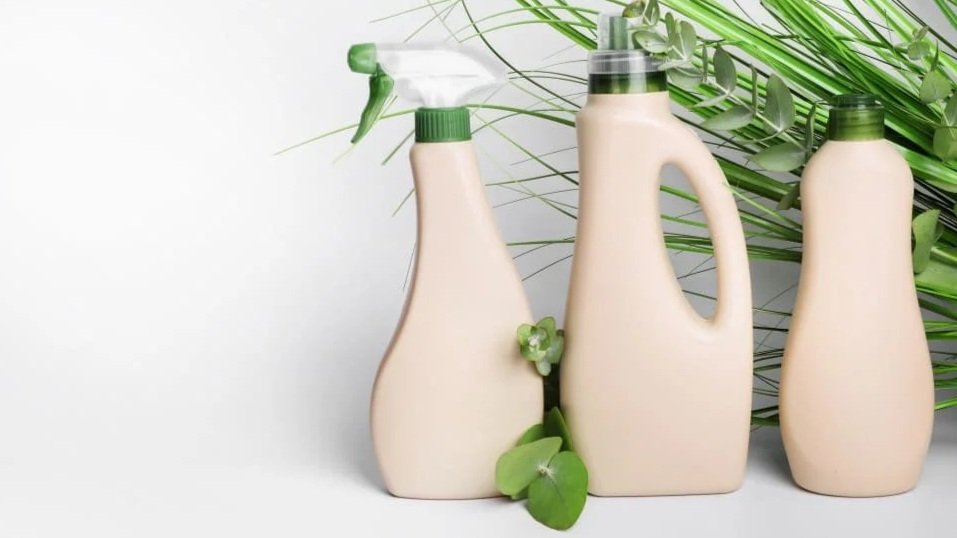 Tre bottiglie di plastica beige con tappi verdi e una pianta verde con foglie vicino, su sfondo bianco con foglioline verdi