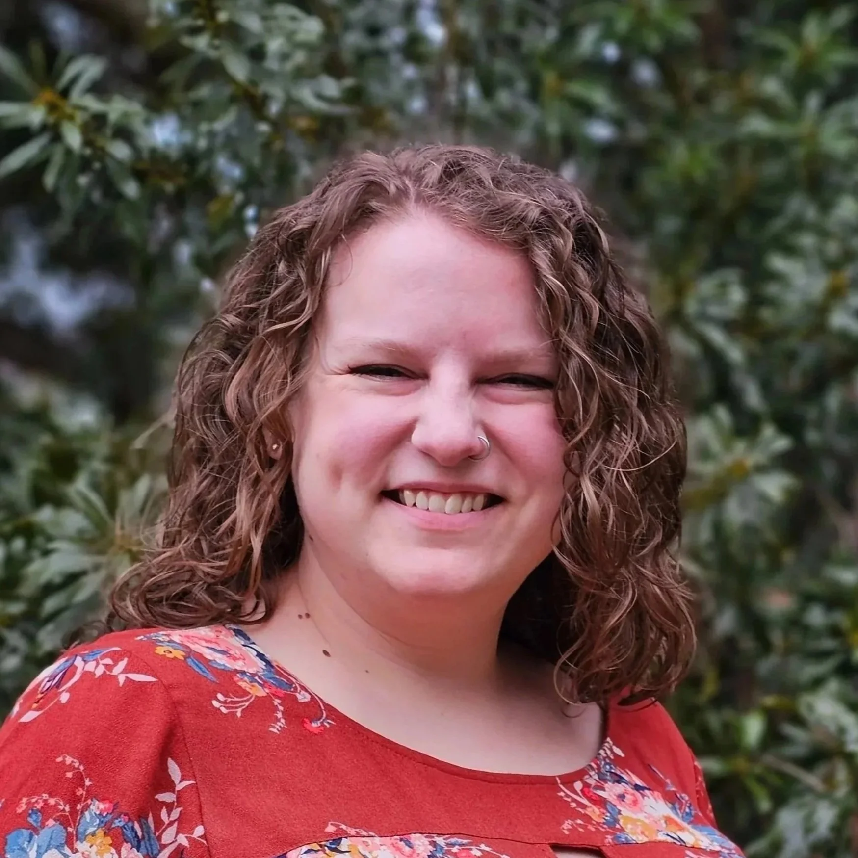 A woman with curly brown hair wearing a red floral top smiling outdoors with trees in the background.