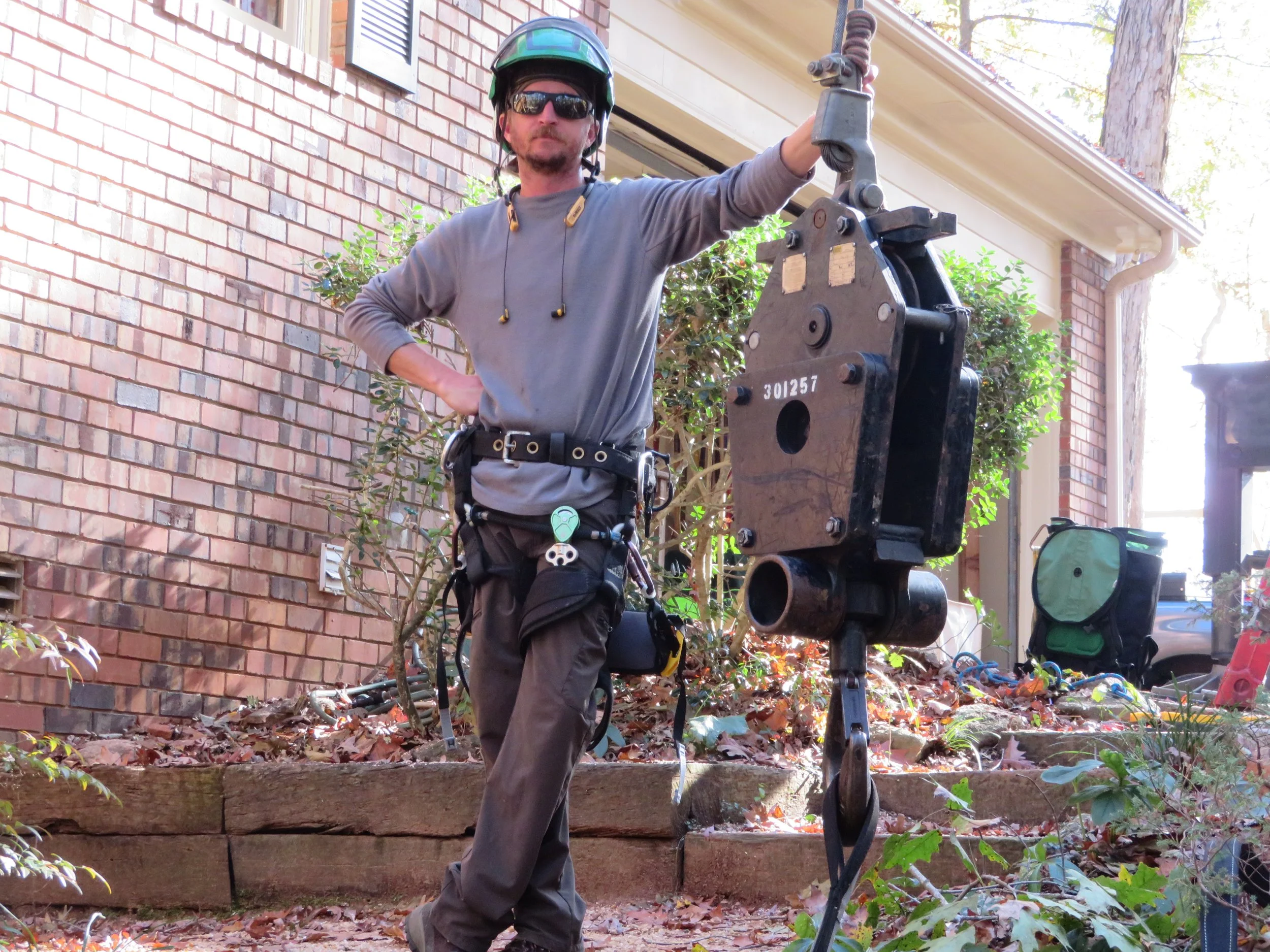 A man standing beside a piece of machinery outdoors, wearing sunglasses, a helmet, and a harness, with one hand on his hip and the other on the machinery.