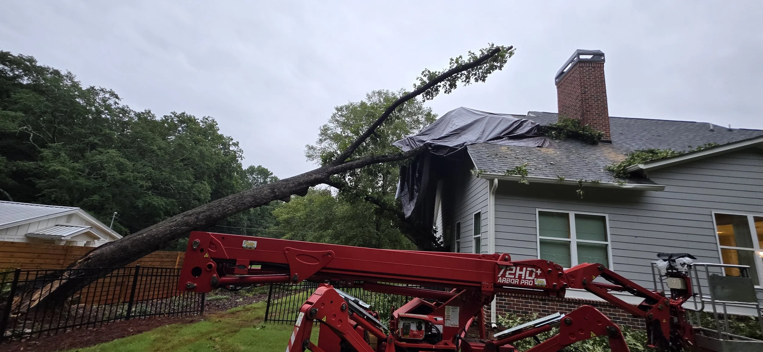 A large fallen tree branch on a house's roof, with storm damage tarp partially covering the roof, and a red cherry picker lift in the foreground. The scene suggests a recent storm or wind damage.