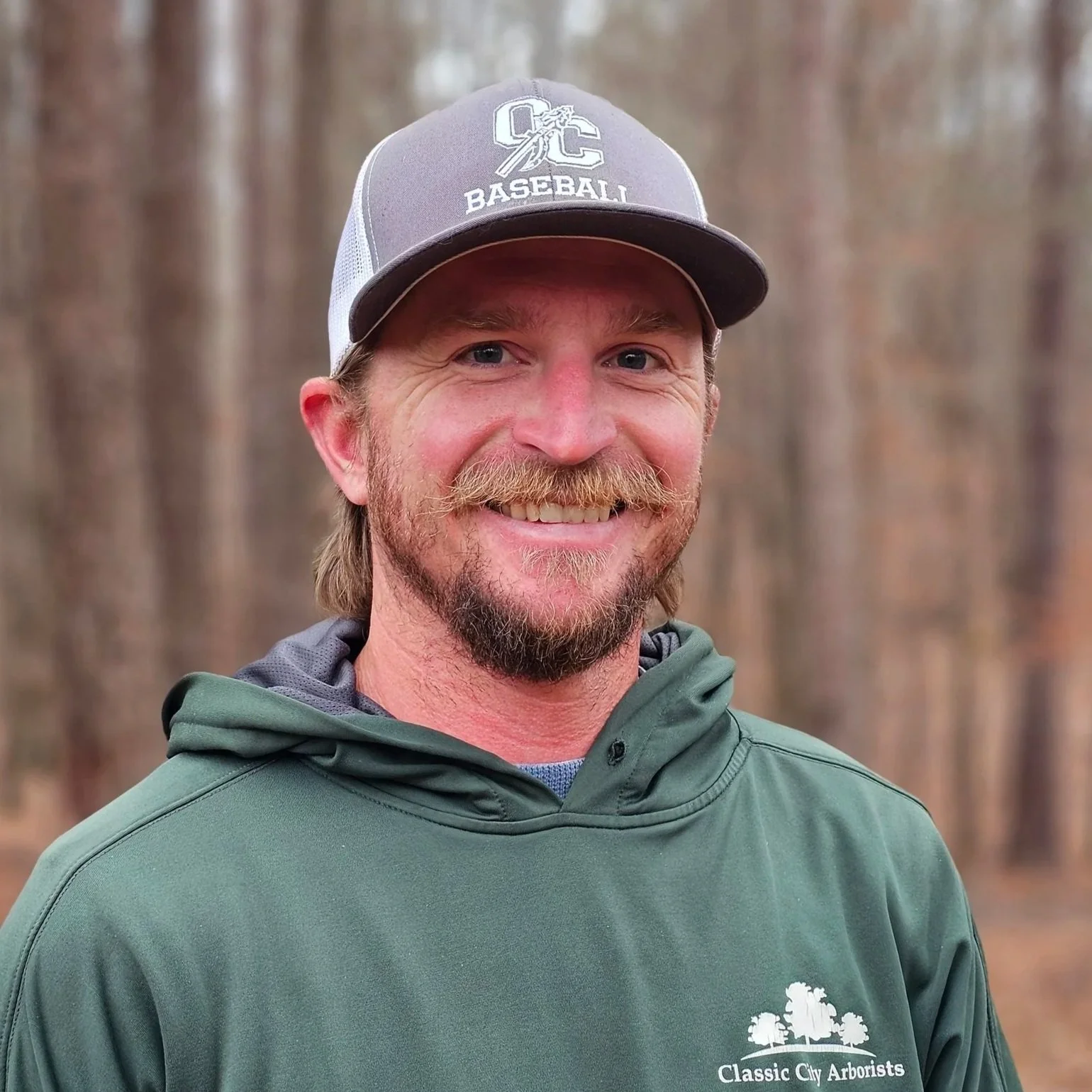 A smiling man with a beard and mustache, wearing a baseball cap and a green hoodie. The cap has a logo that reads "QC Baseball," and the hoodie features the logo of Classic City Arborists. He is outdoors with a blurred background of trees.