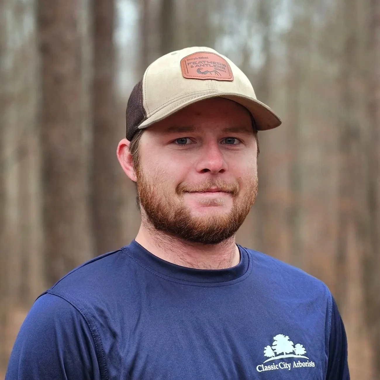 A man with a beard and blue eyes wearing a tan baseball cap and a navy blue shirt with a logo of a tree and the text "Classic City Arborists". He stands outdoors with blurred trees in the background.