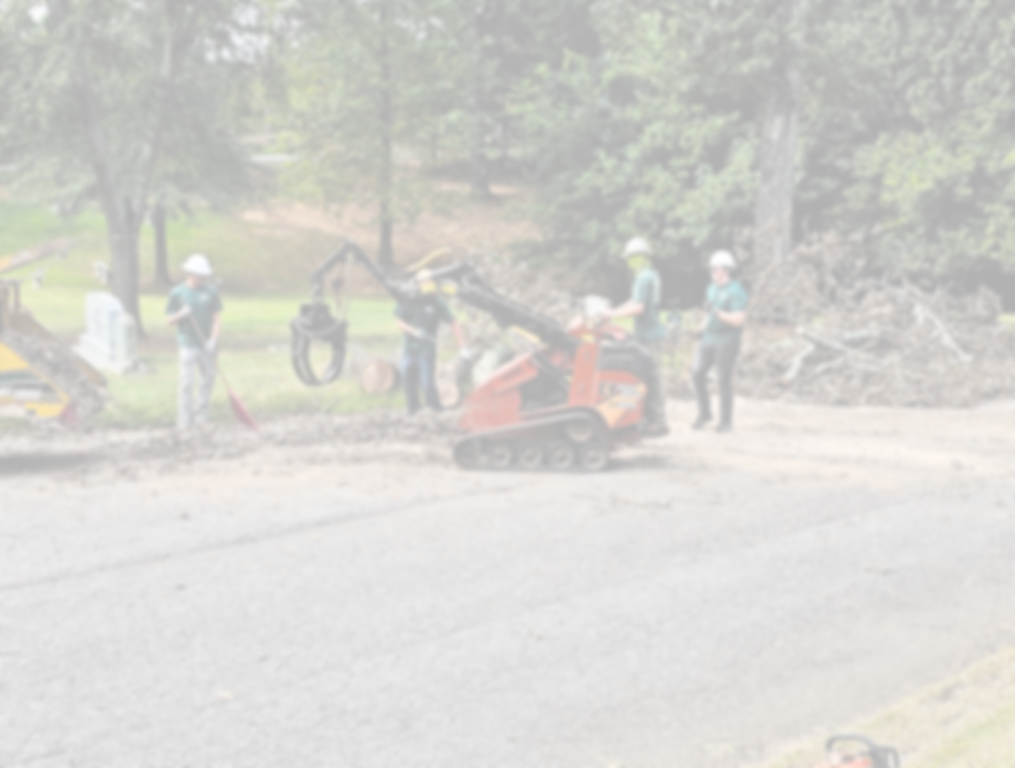 Workers in safety helmets operate and monitor landscaping machinery and tools on a construction site surrounded by trees.