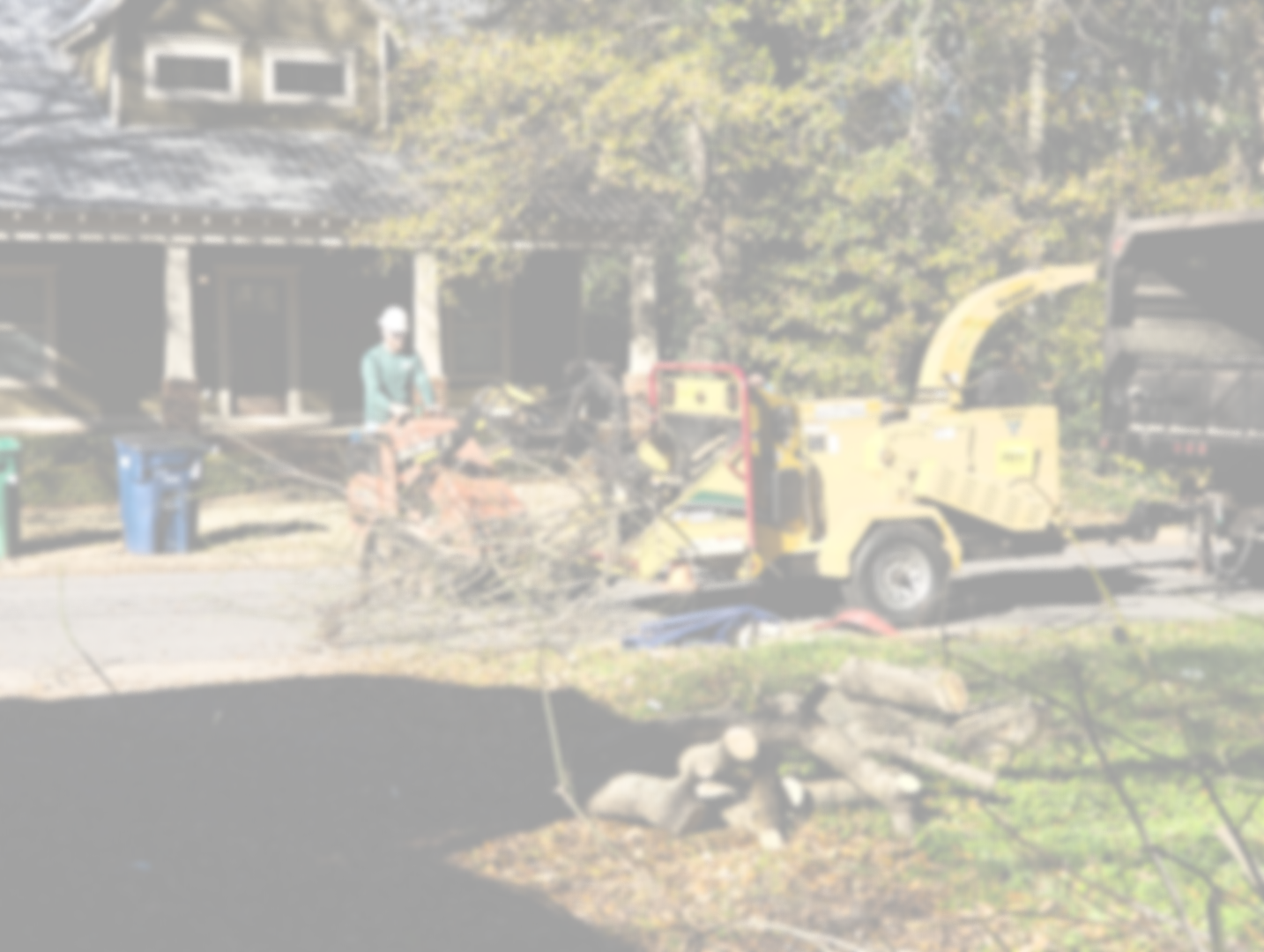 A man operating a wood chipper on a residential street in front of a house, with a truck parked nearby and logs on the ground.