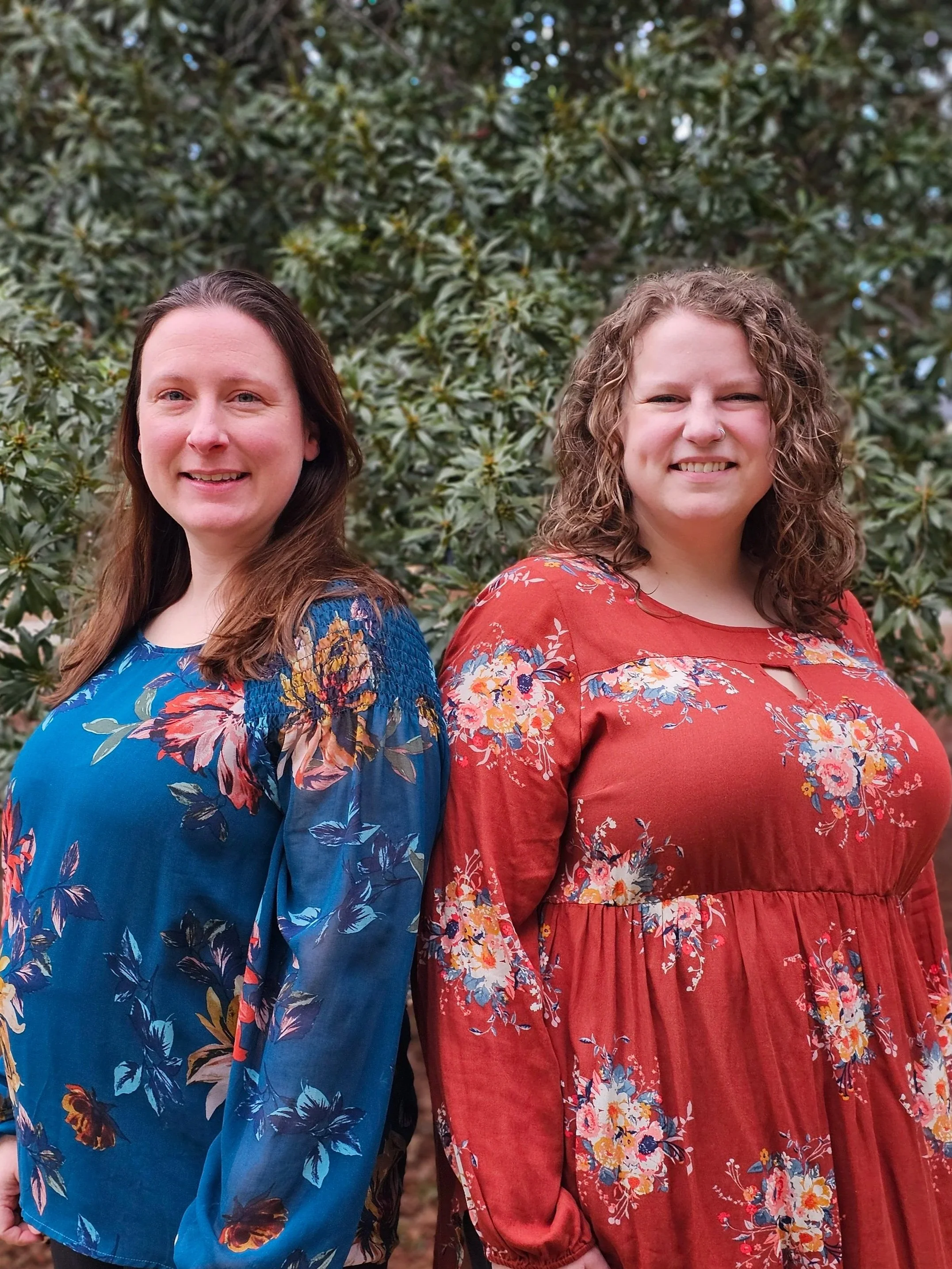 Two women standing outdoors in front of green foliage, smiling, wearing colorful floral dresses.