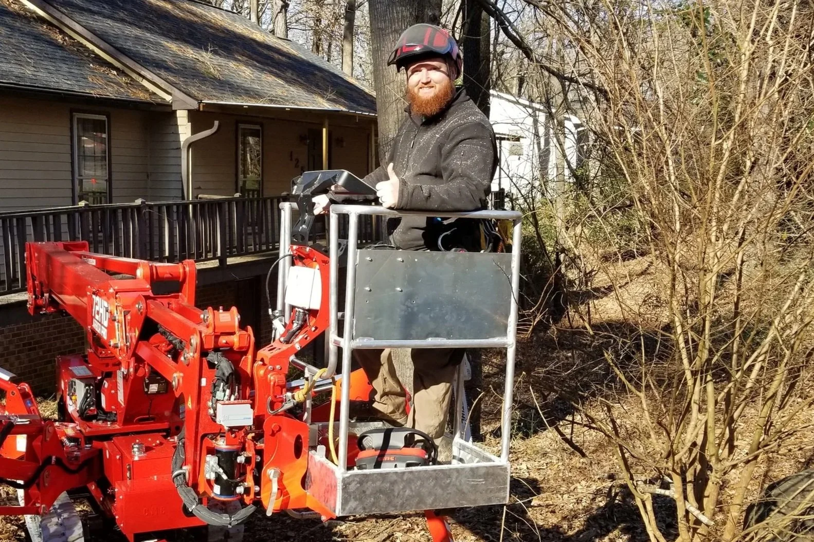 A man with a beard wearing a helmet and gloves standing on a cherry picker lift, giving a thumbs-up, in a backyard with trees and a house in the background.