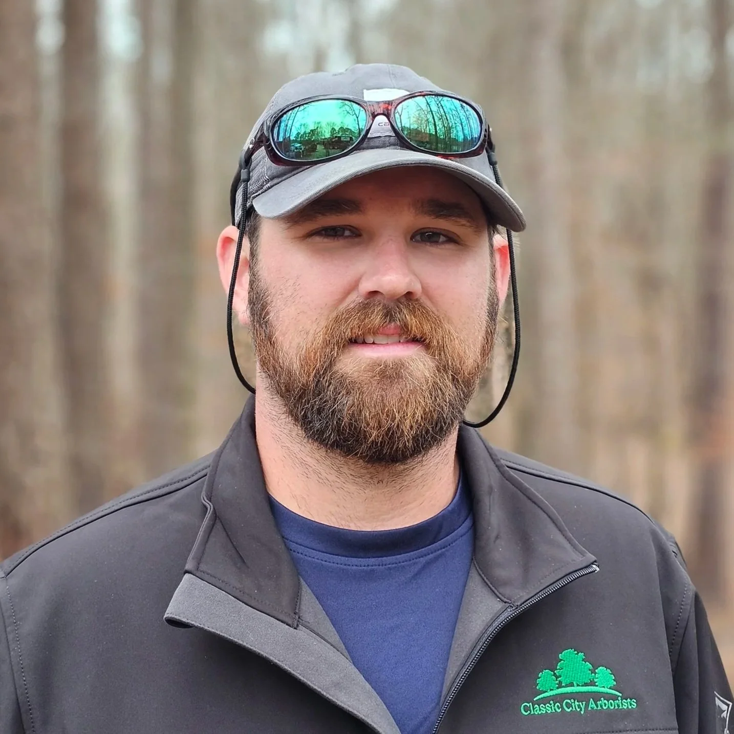 Portrait of a man with a beard wearing a gray cap with sunglasses on top, a jacket with a logo that says 'Classic City Arborists', and a forest background.