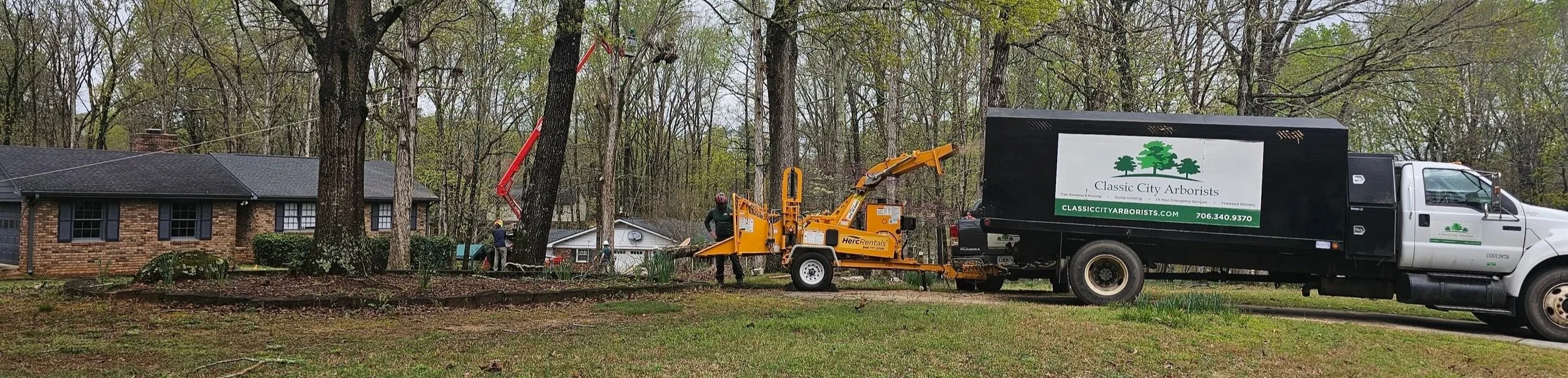 Tree trimming truck operated by Classic City Arborists trimming trees in a residential yard with workers and trees visible, house and wooded area in the background.