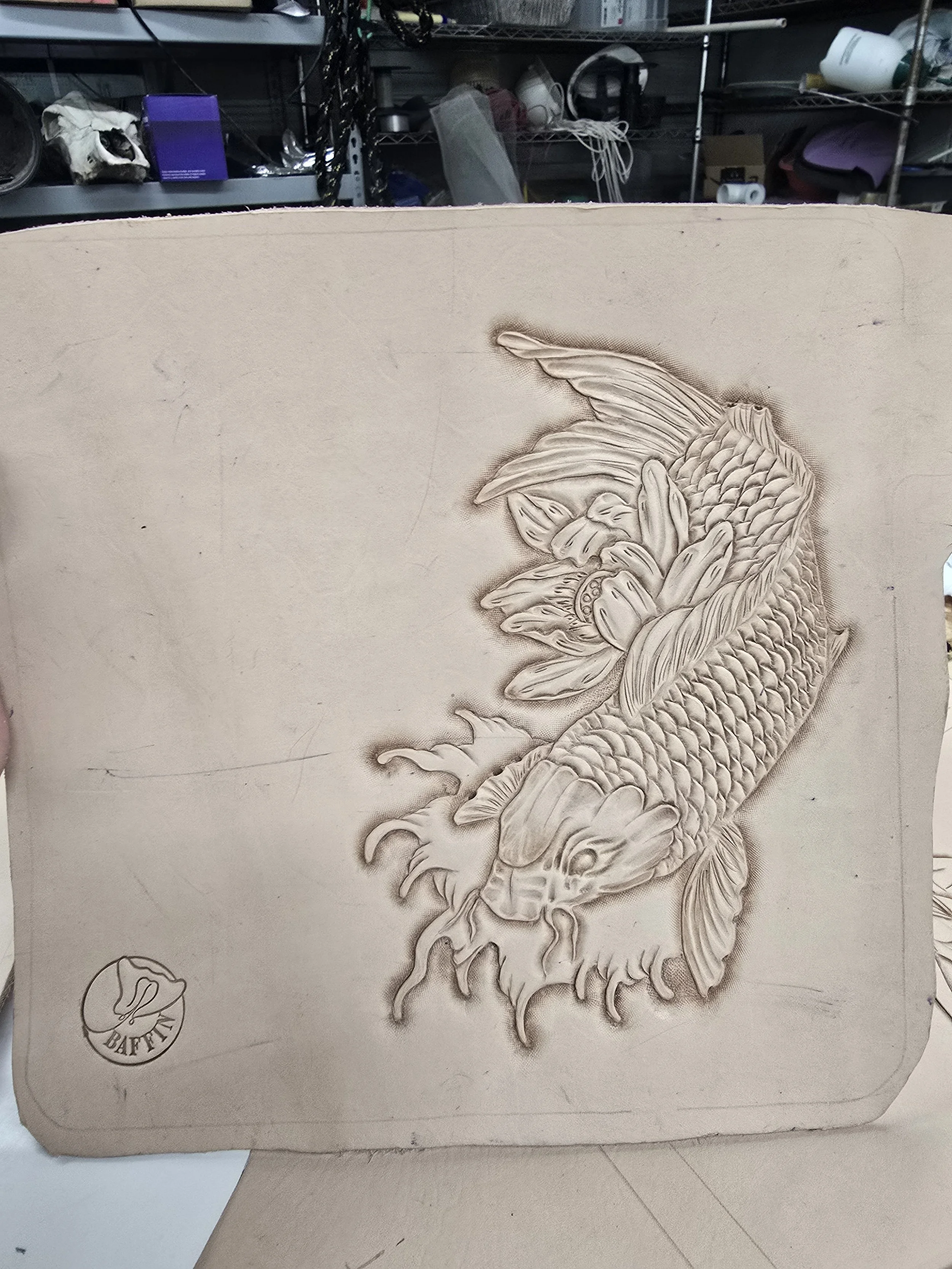 Unfinished beige ceramic tile with an embossed koi fish design, showing detailed scales, fins, and flowing water around its body, with a small logo in the bottom left corner.